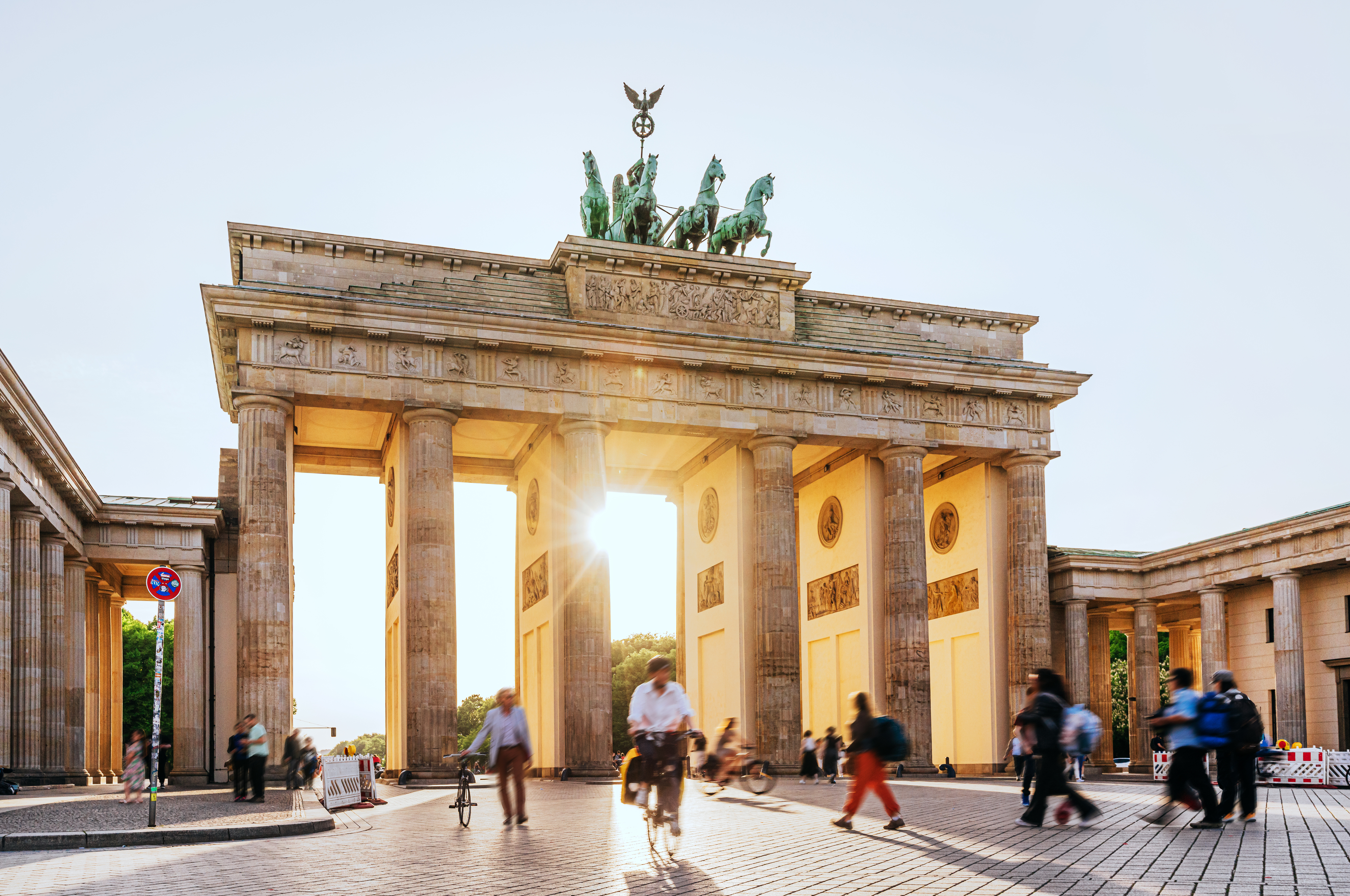 People walking and cycling near the Brandenburg Gate in Berlin as sunlight streams through its columns