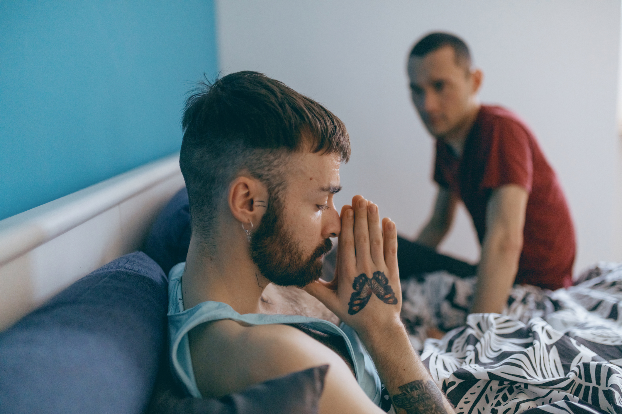 Two men, one with a tattoo on his hand, sit pensively in a bedroom on a bed covered with a patterned blanket