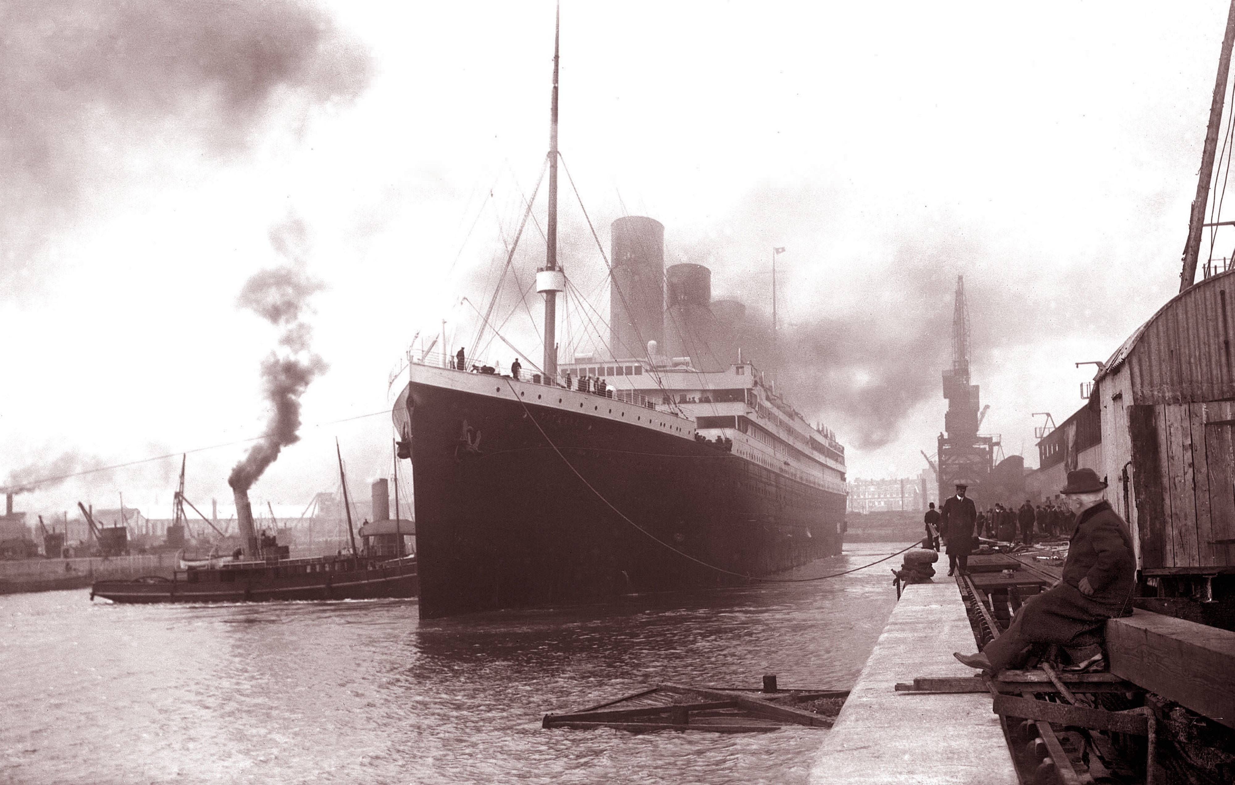 Vintage photograph of the RMS Titanic docked at a shipyard with smoke billowing from its funnels, and workers seen on the dock and nearby buildings