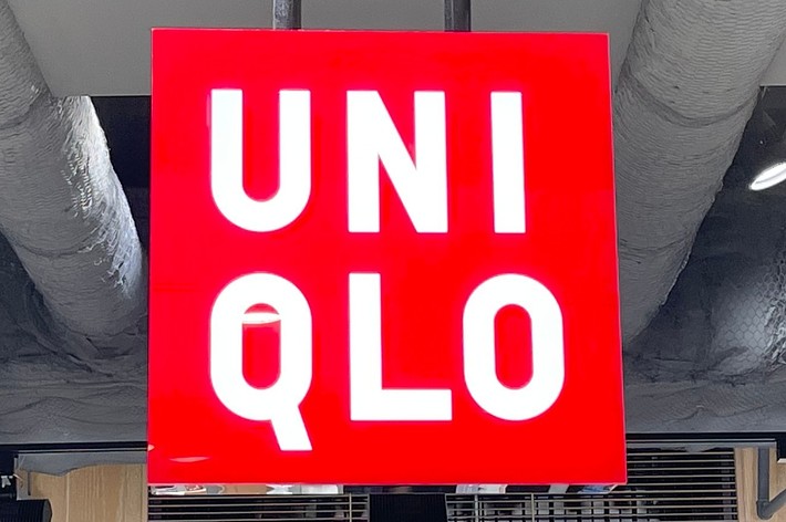 Uniqlo store interior with a prominent red and white sign reading “Uniqlo” hanging from the ceiling. Shelves with clothing are visible below the sign