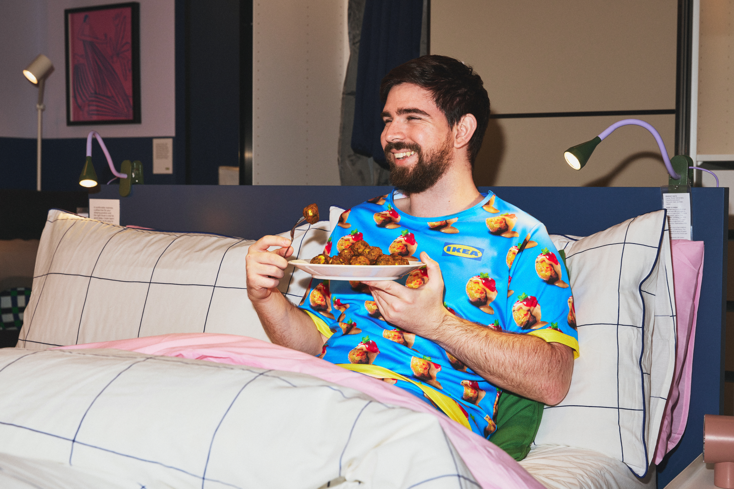 Man dressed in a shirt with a meatball pattern enjoying food in bed with modern decor in the background