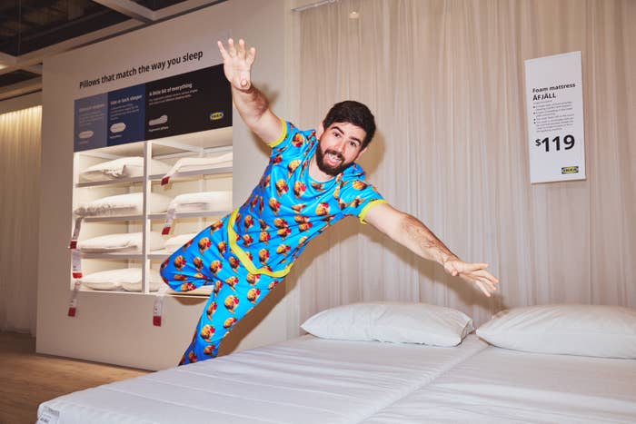 Man in playful pajama set dramatically falling onto a white bed in a showroom, showcasing pillows and mattresses behind him with promotional signs