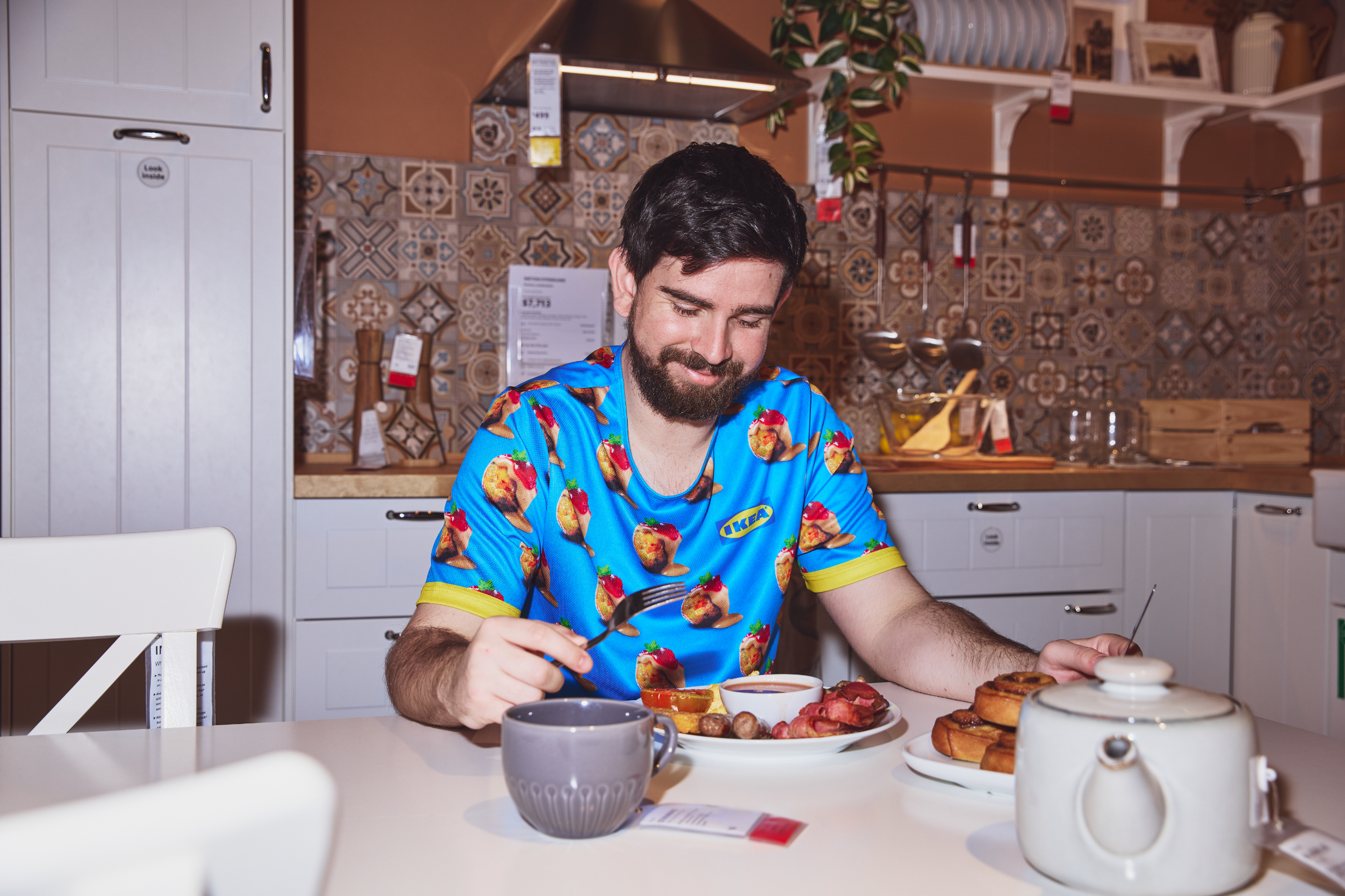 Man in a kitchen wearing a fruit-themed shirt, smiling while having breakfast with pancakes and bacon. Kitchen has a tiled backsplash and various cooking utensils