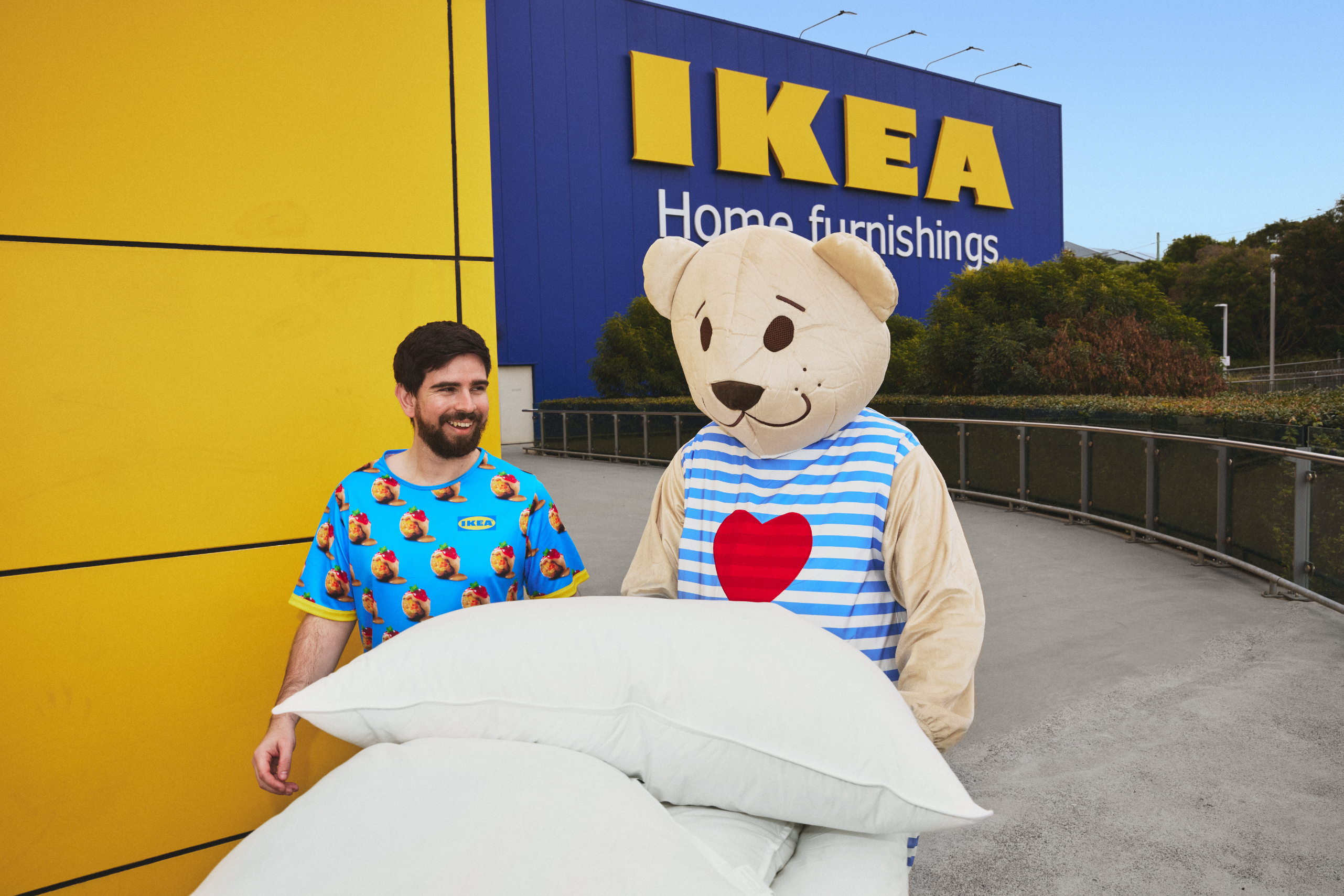 Man in IKEA shirt holding pillows stands with person in teddy bear costume wearing striped shirt with heart, in front of IKEA store