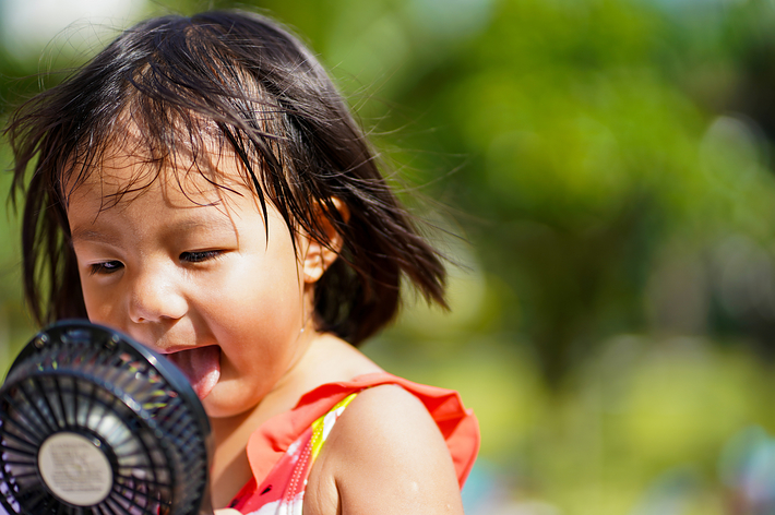 小さな女の子が手持ちの扇風機に顔を近づけている。