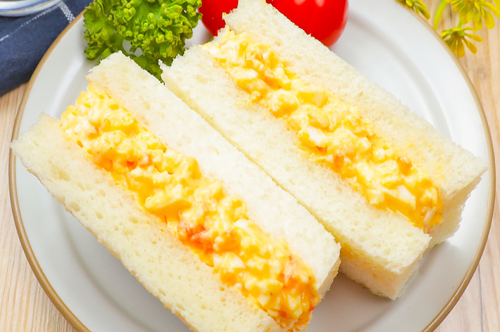 白い皿に盛り付けられた卵サンドイッチとパセリ、チェリートマトの写真。周囲には植物が飾られている。