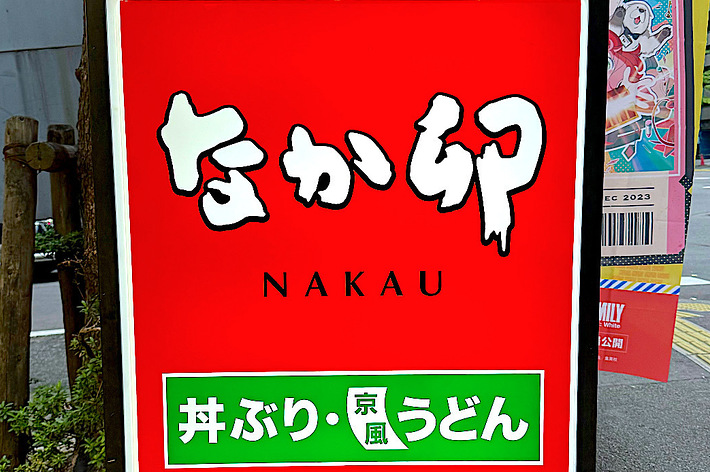 A sign for the Japanese restaurant Nakau promoting their dishes, featuring text in Japanese and English, located on a city sidewalk
