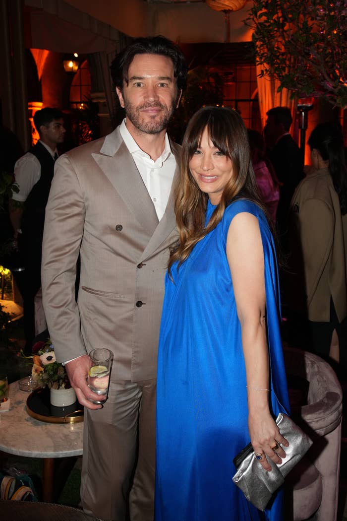 Tom Pelphrey and Kaley Cuoco are standing together at an event. Tom is wearing a suit, and Kaley is in a sleeveless dress, holding a clutch