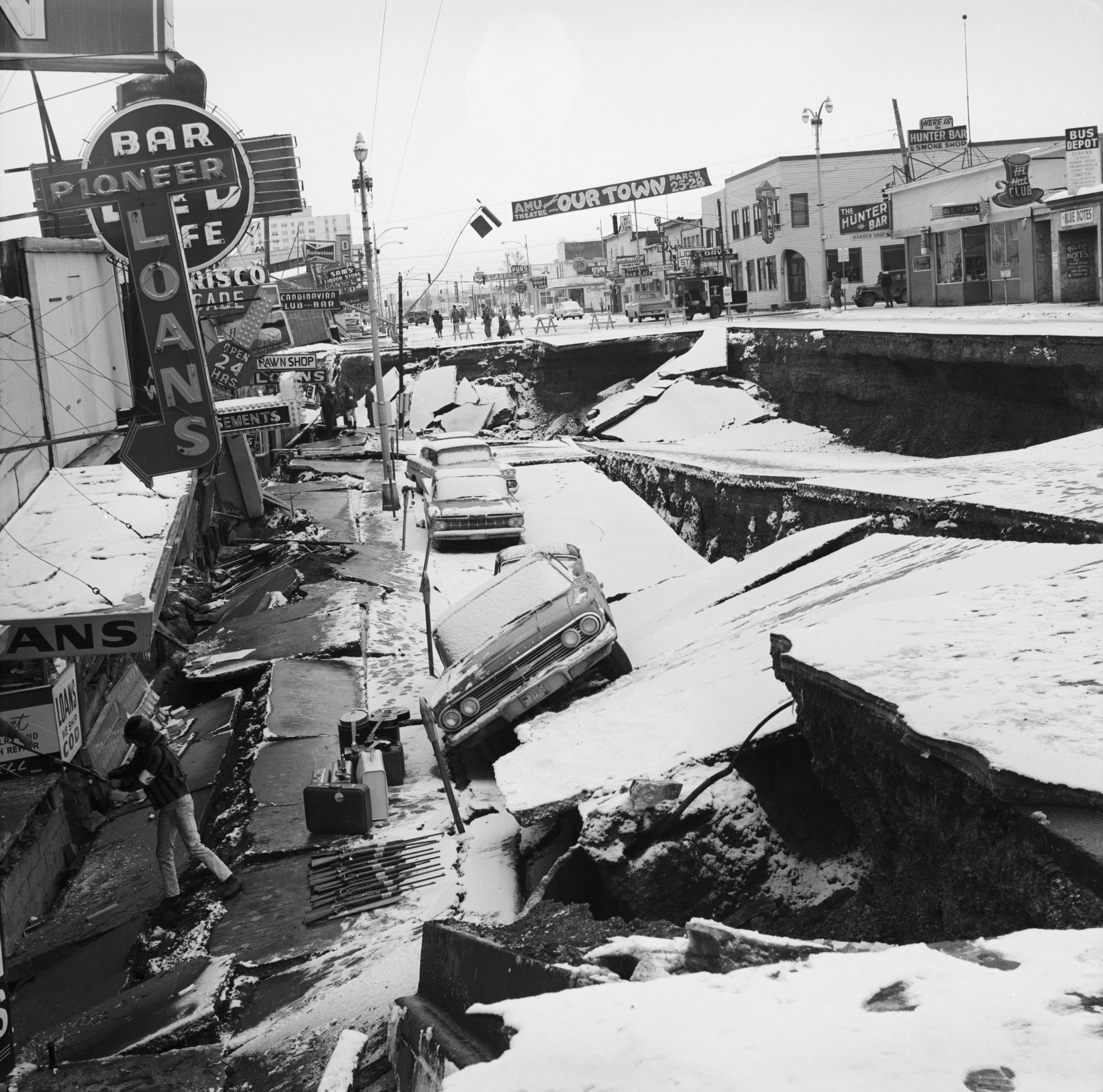 Street scene of an earthquake in Anchorage, Alaska, 1964, showing damaged roads, toppled cars, and disrupted buildings with various business signs visible