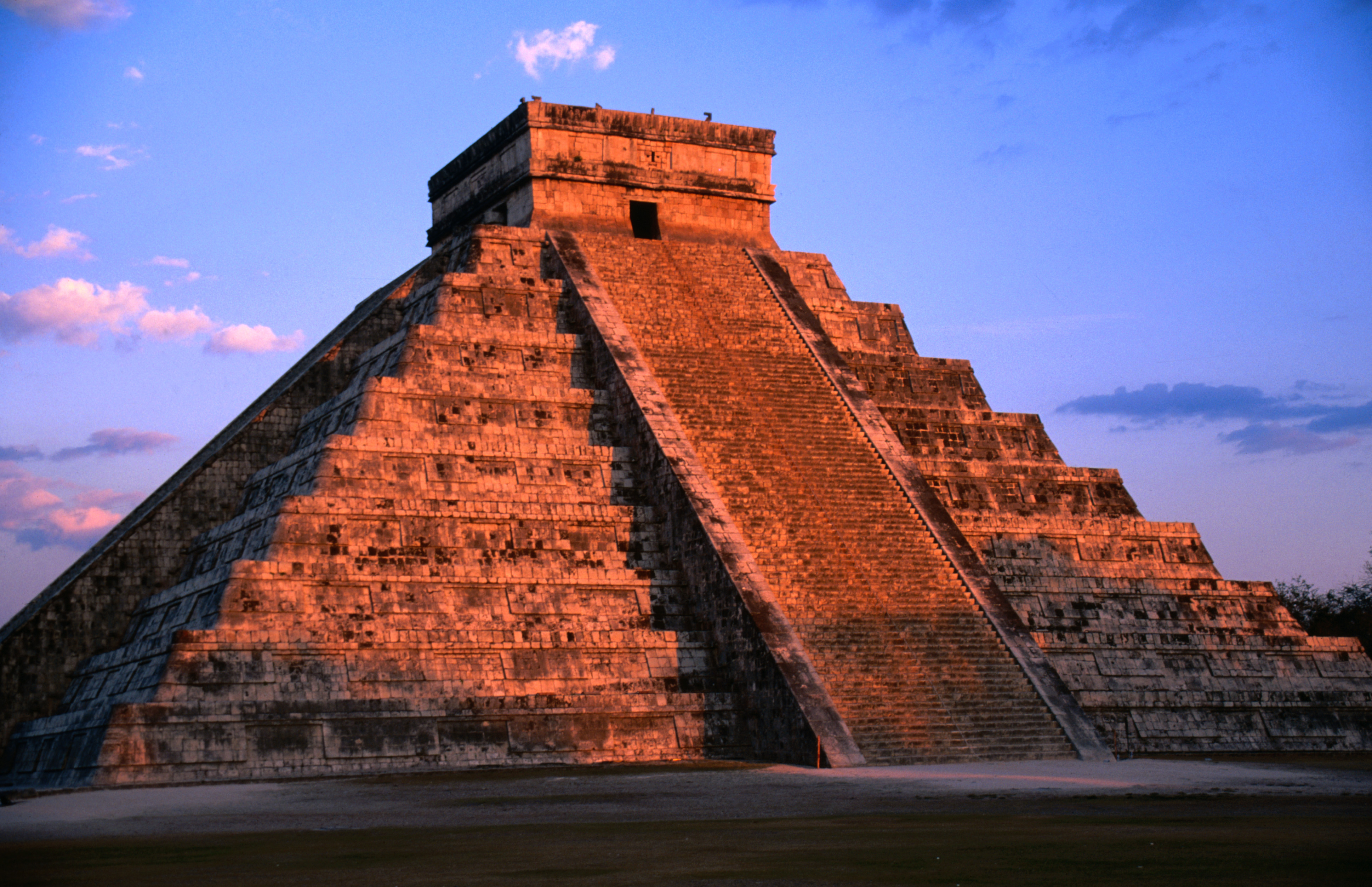 El Castillo, the pyramid at Chichen Itza, stands under a partly cloudy sky at sunset. The staircase and structure are prominently visible