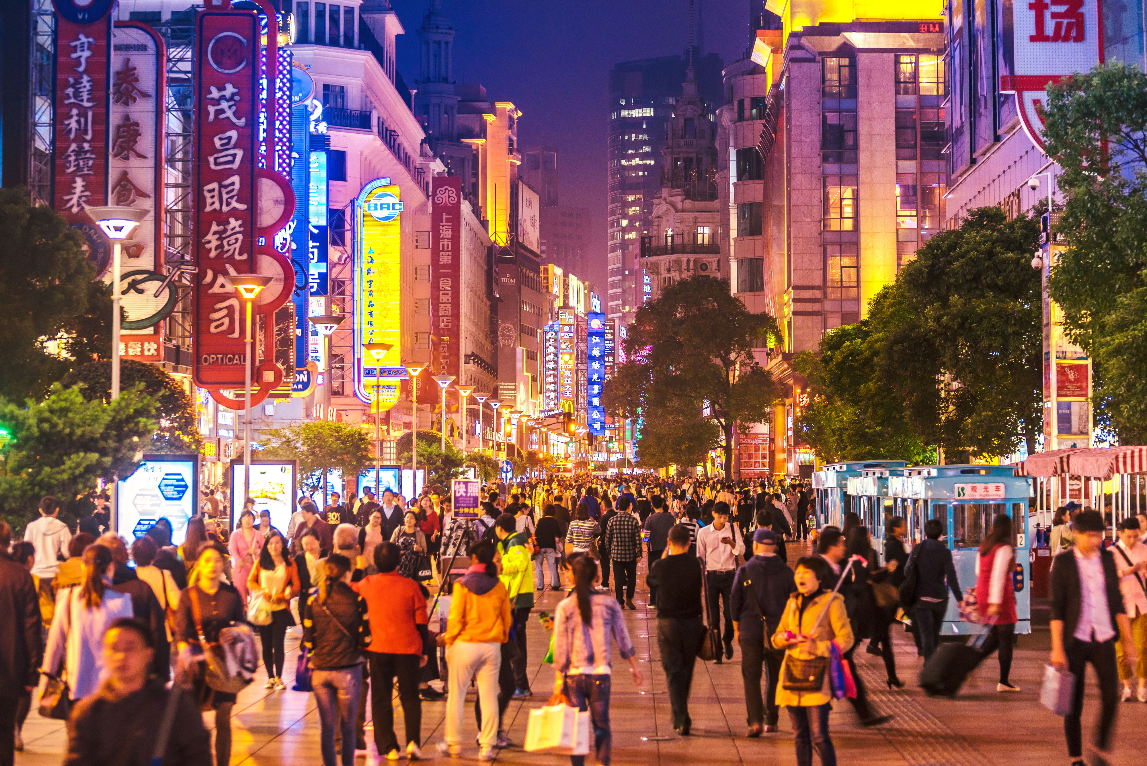 A busy commercial street in an urban area at night with many illuminated signs and people walking on the sidewalk