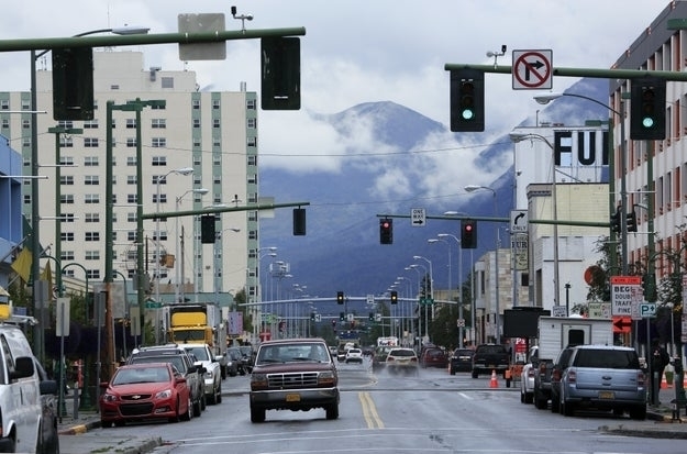 Busy city street with traffic and a mountainous backdrop. Various vehicles, traffic lights, and buildings are visible