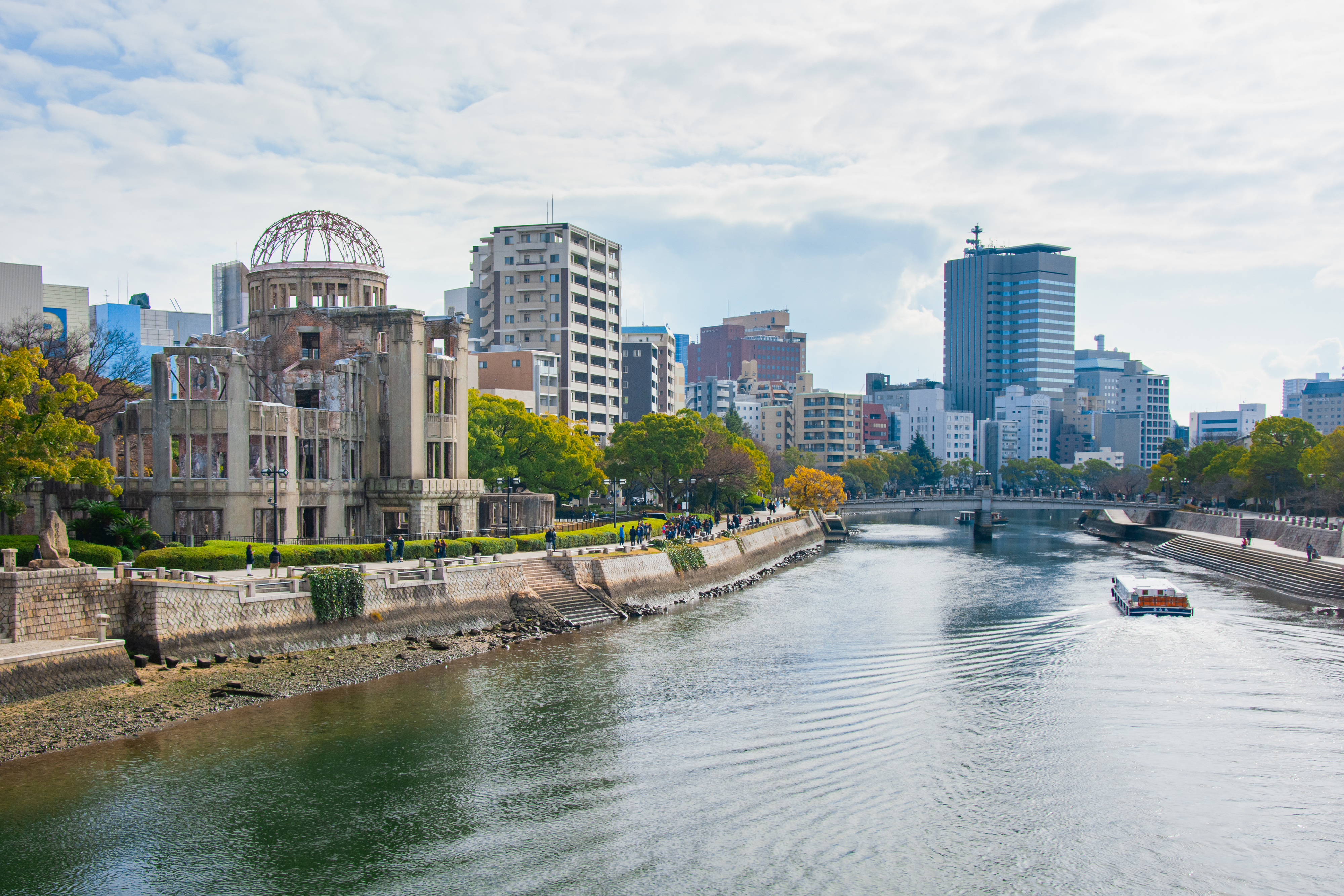 A daytime view of Hiroshima Peace Memorial, with city buildings in the background and a river with a boat in the foreground