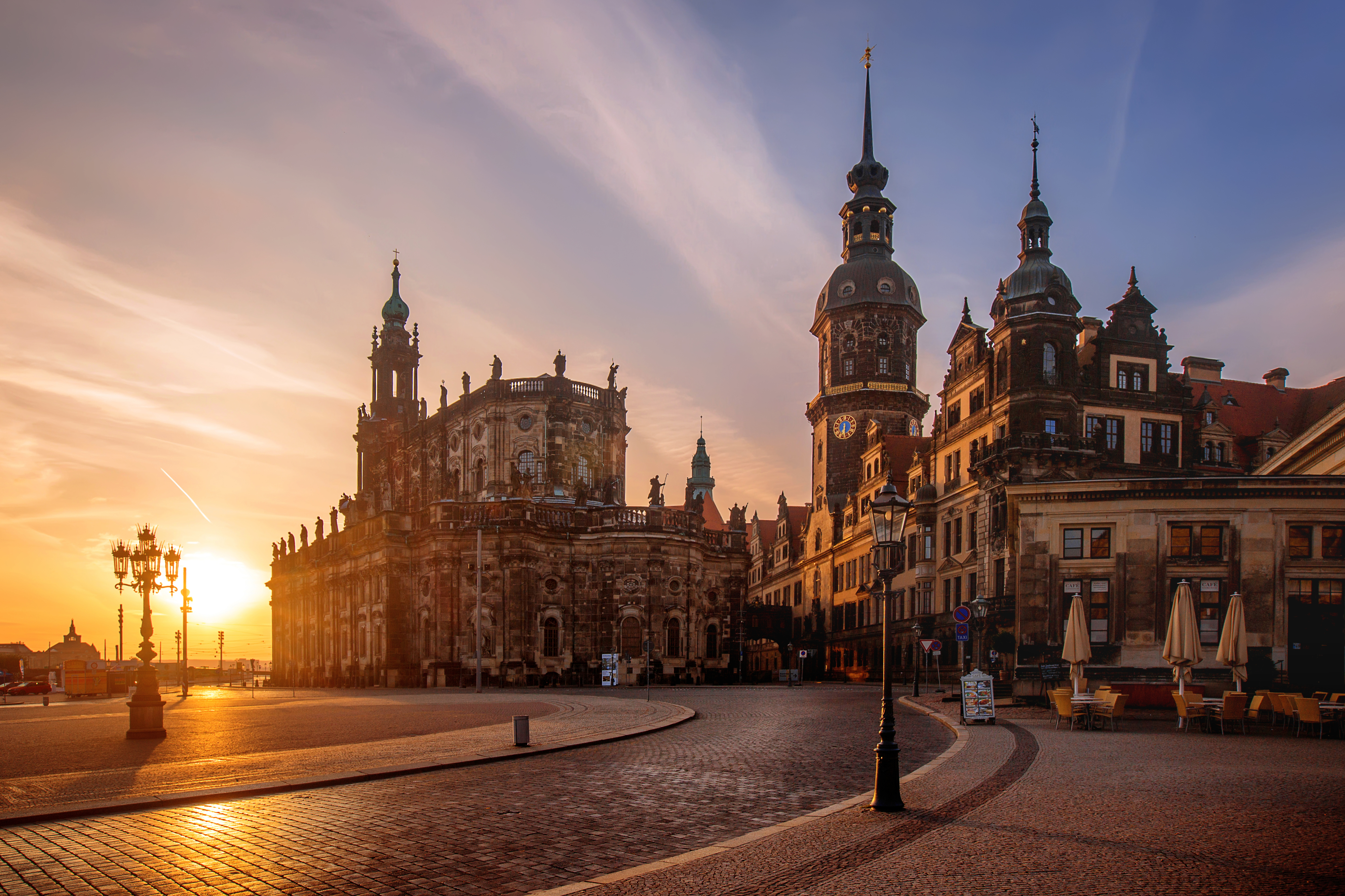 Historic European plaza at sunrise, featuring an ornate cathedral with a tall clock tower and surrounding classical architecture. Few people are present