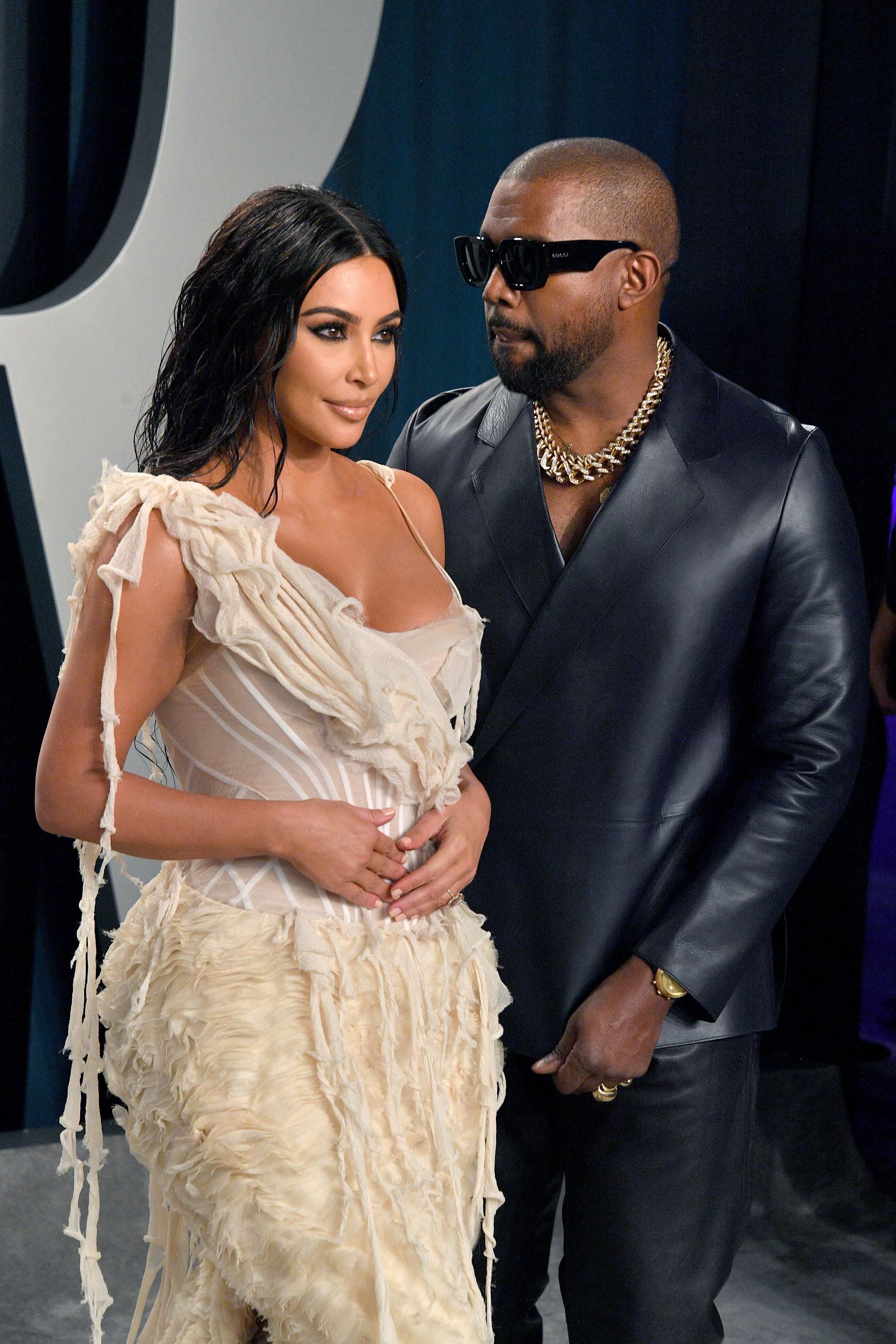 Kim Kardashian in a feathered, detailed dress and Kanye West in a dark leather jacket with a gold chain necklace pose at an event