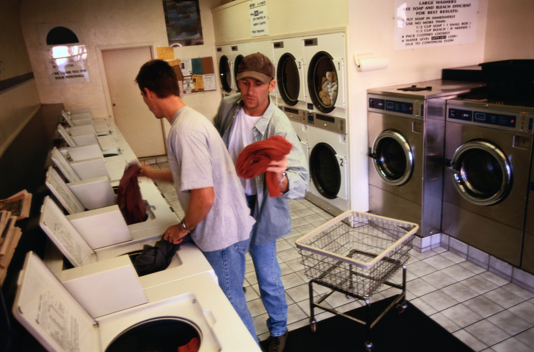Two men doing laundry in a laundromat, one putting clothes into a washing machine, the other holding a garment