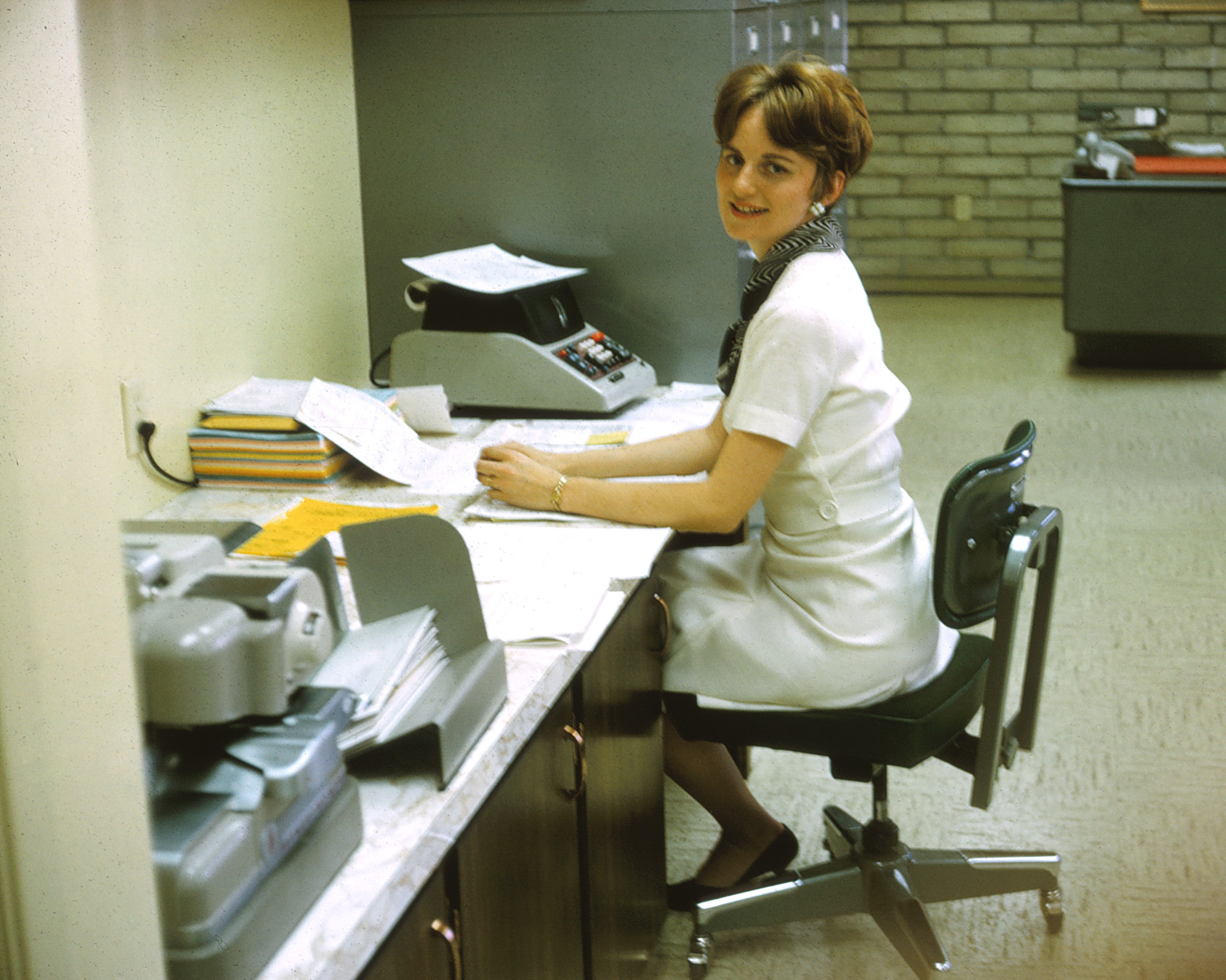 Smiling woman in 1960s office attire, seated at a desk with papers, books, and office equipment, looking at the camera