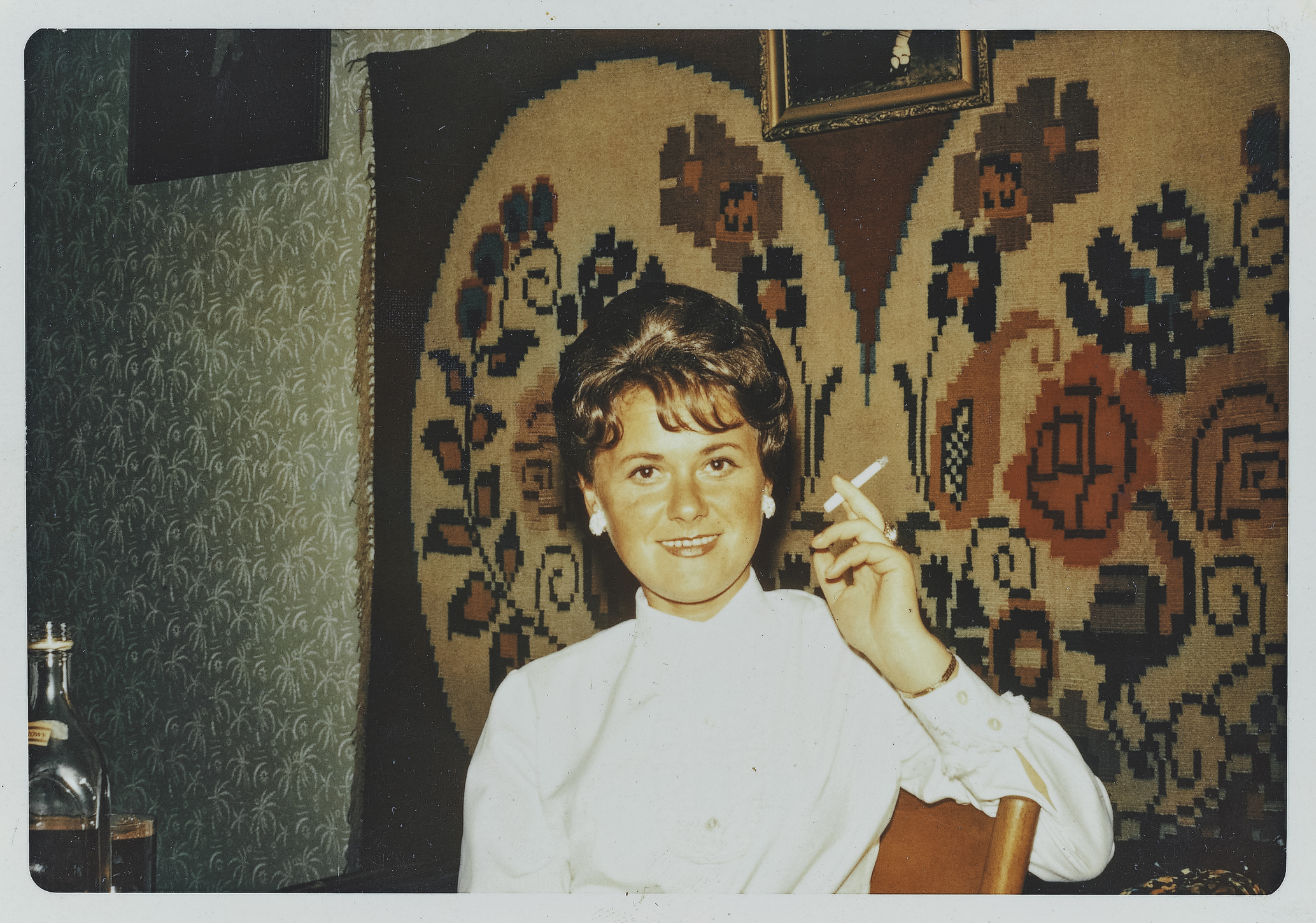 A woman in a white blouse sits in a room with a floral tapestry, holding a cigarette and smiling