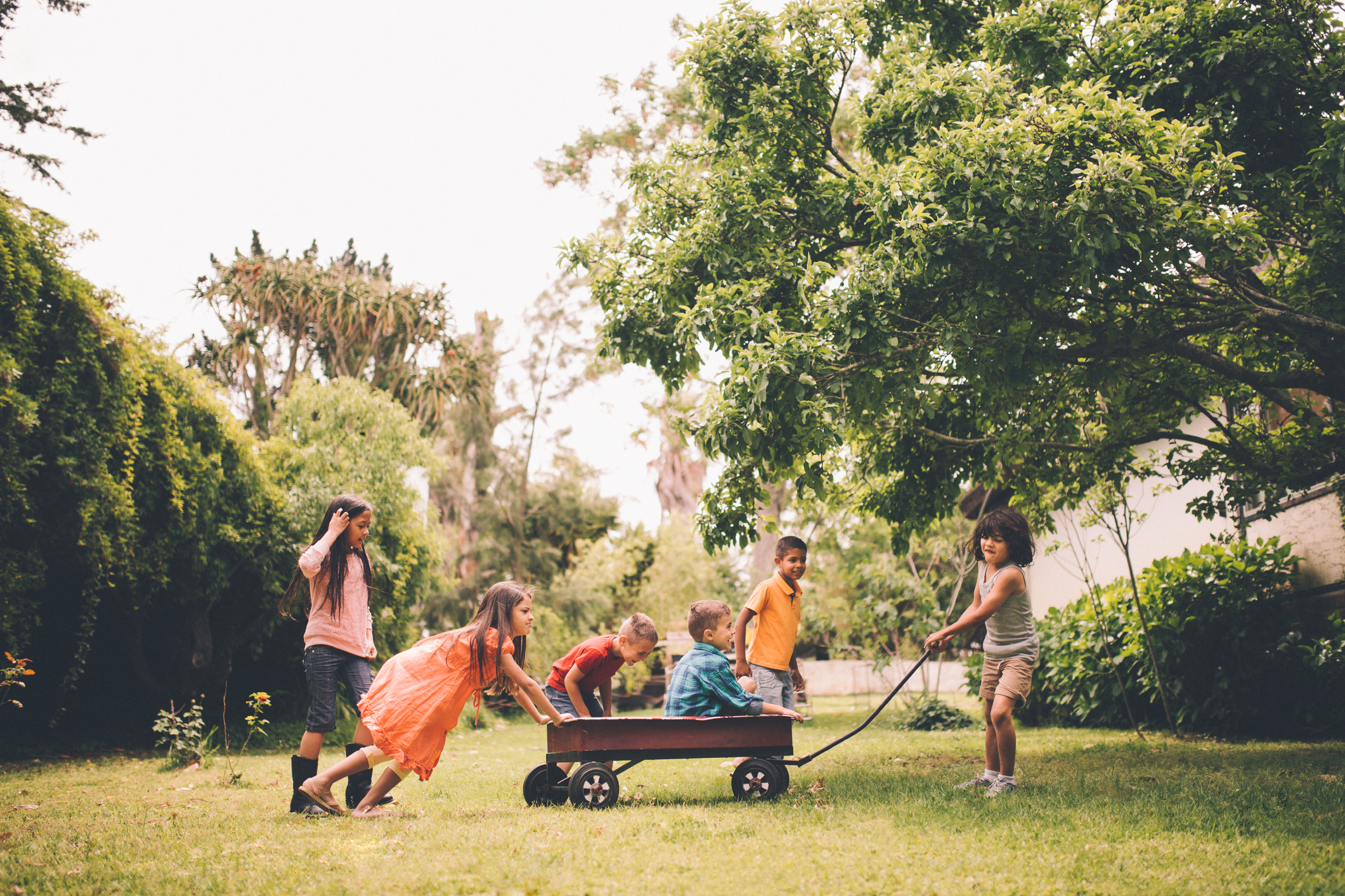 Children are playing outside, with five kids around a wagon in a grassy yard. One child pulls the wagon while two others push, and two kids are inside the wagon