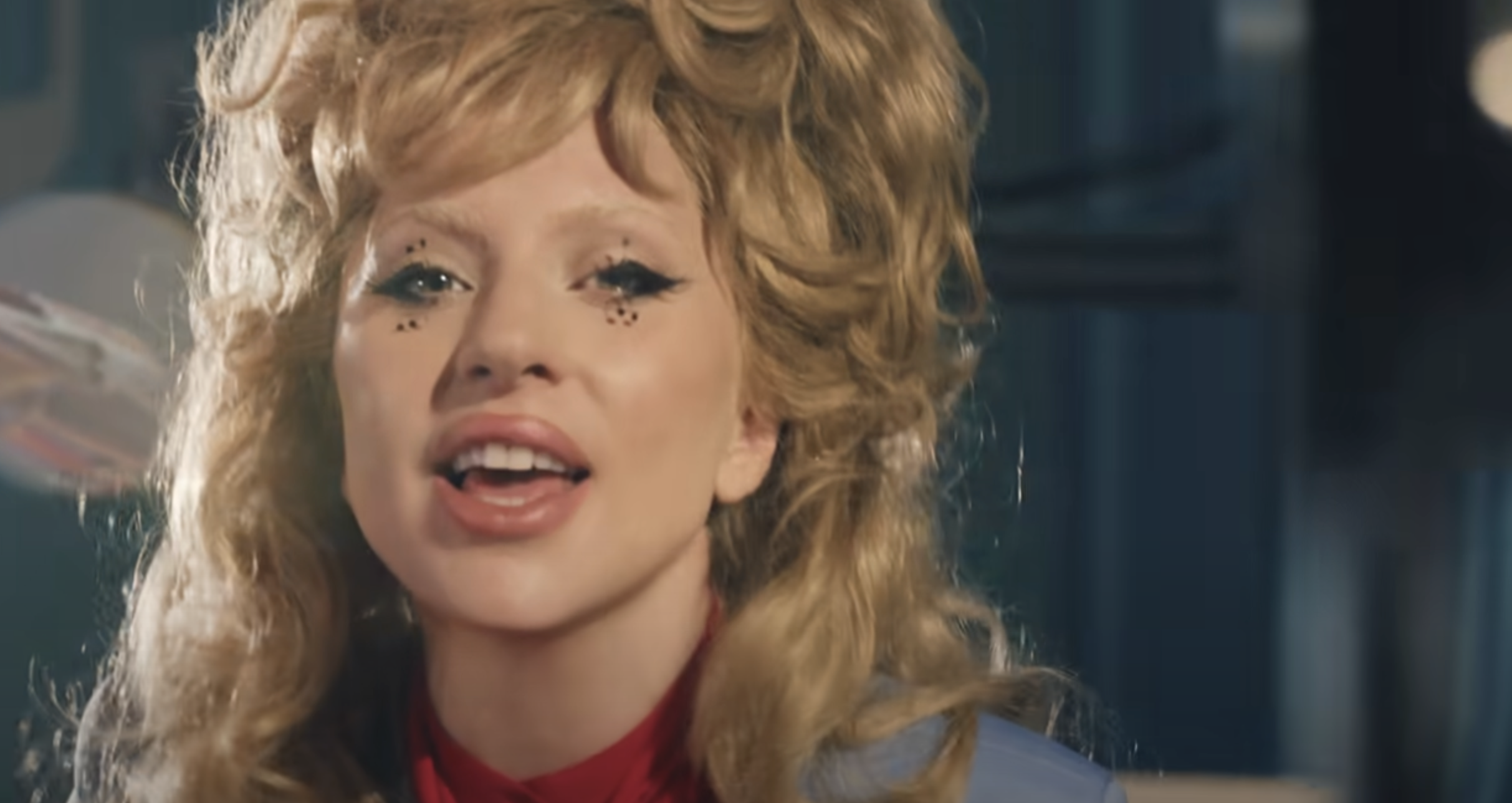 A person with voluminous hair and star-like makeup details around their eyes, seen performing or speaking