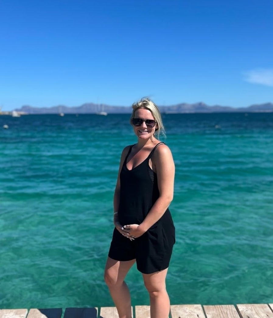 Pregnant person in a black romper stands on a wooden dock by the ocean, smiling with sunglasses on