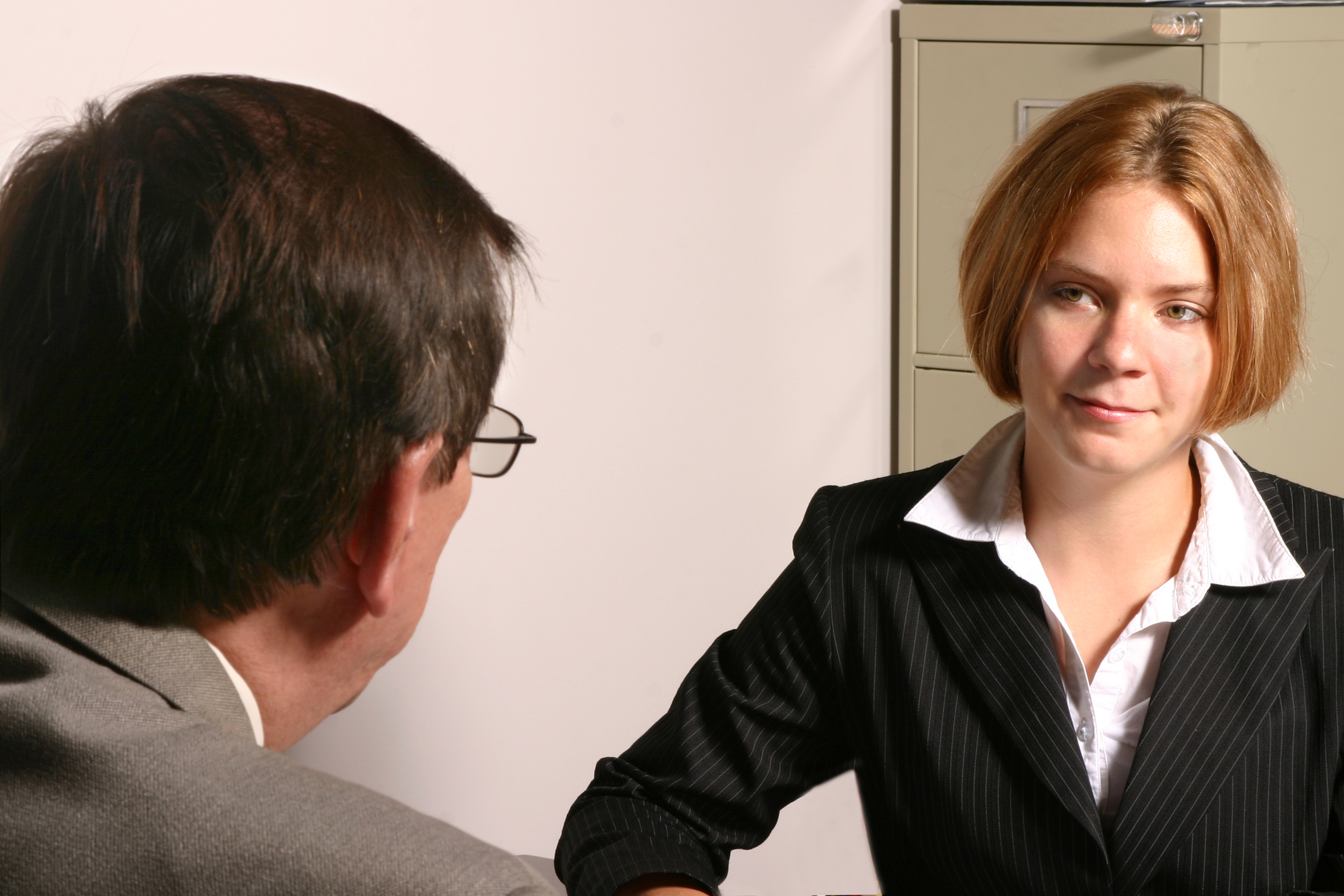 A woman in a business suit is talking to a man in an office setting with a filing cabinet in the background. Their expressions are neutral