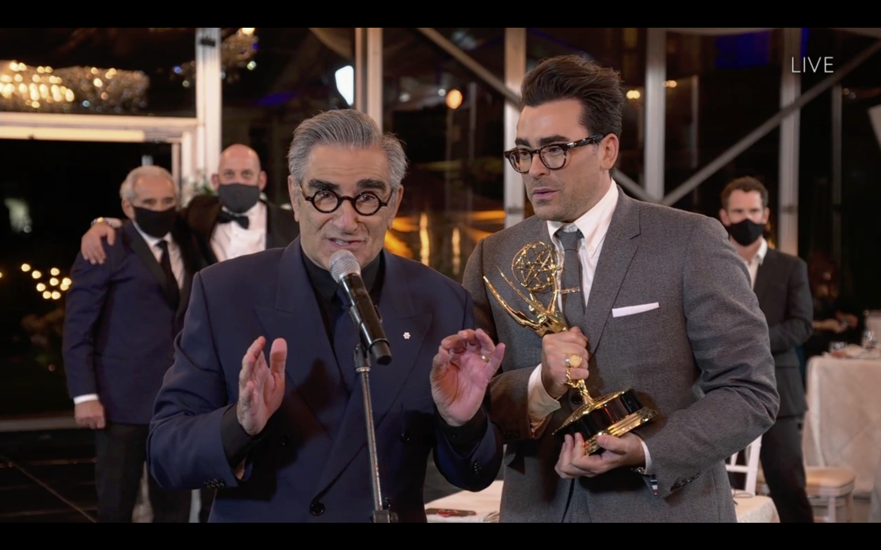 Eugene Levy and Dan Levy stand at a microphone, with Dan holding an Emmy. Both are in formalwear, and two masked men are in the background