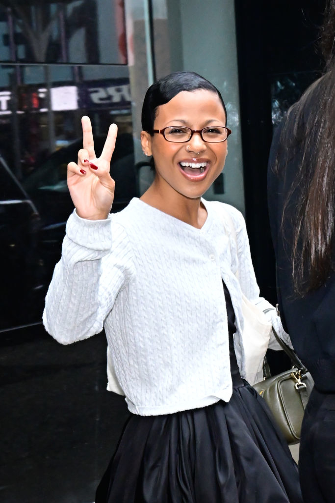 Smiling woman with glasses and an updo, wearing a white textured cardigan over a black dress, gives a peace sign