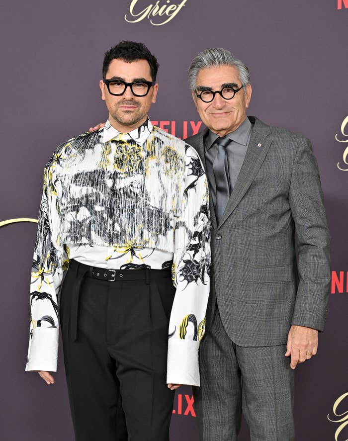 Dan Levy and Eugene Levy standing together at a Netflix event, both wearing glasses. Dan is in a patterned top and black trousers, Eugene is in a gray suit