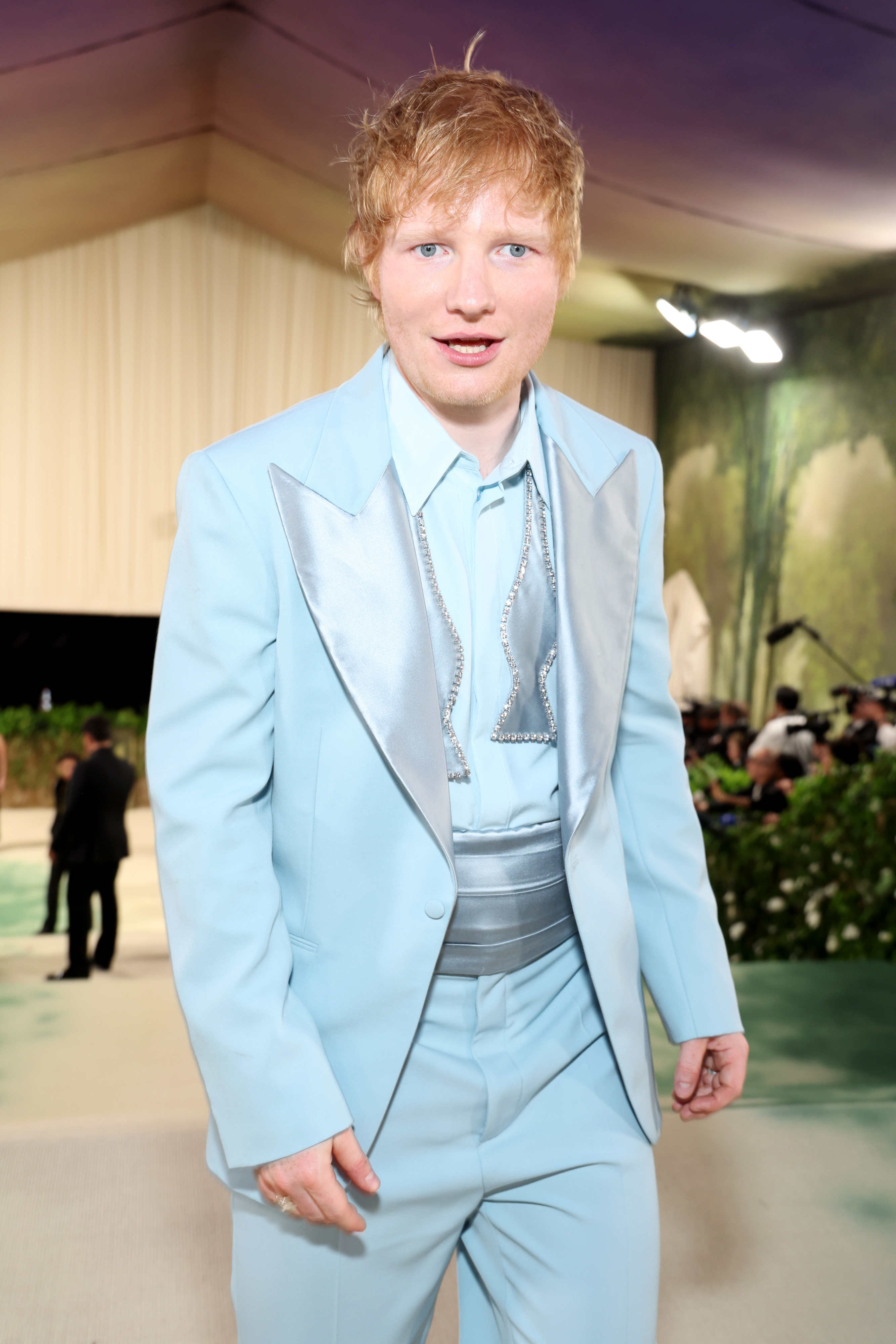 Ed Sheeran at a formal event, wearing a light-colored suit with a metallic tie and a wide waistband