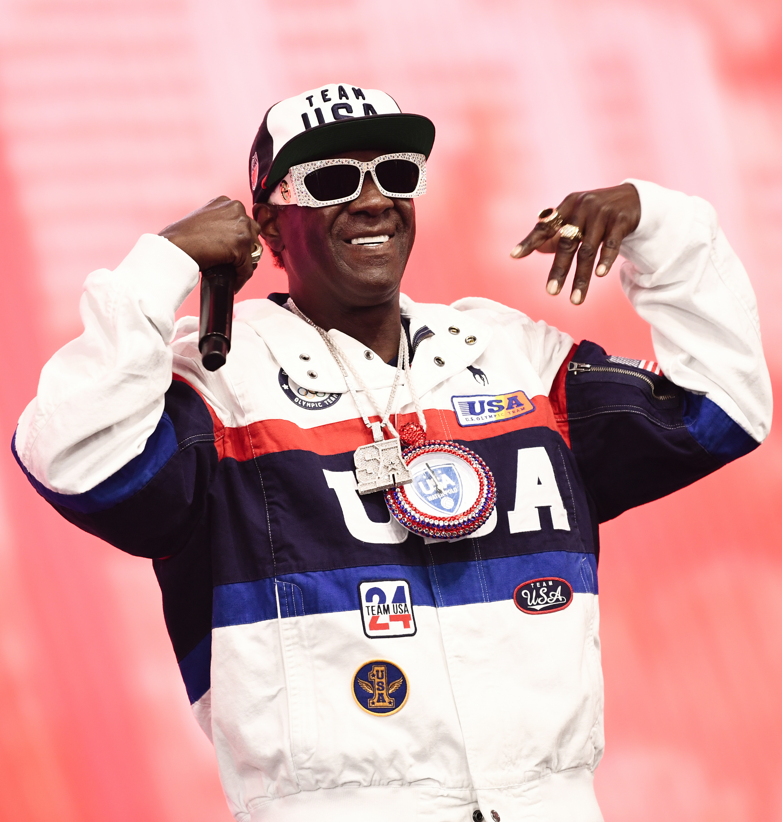 Flavor Flav wears a Team USA jacket, a hat, sunglasses, and a large necklace medallion, posing with his hands up, at a public event