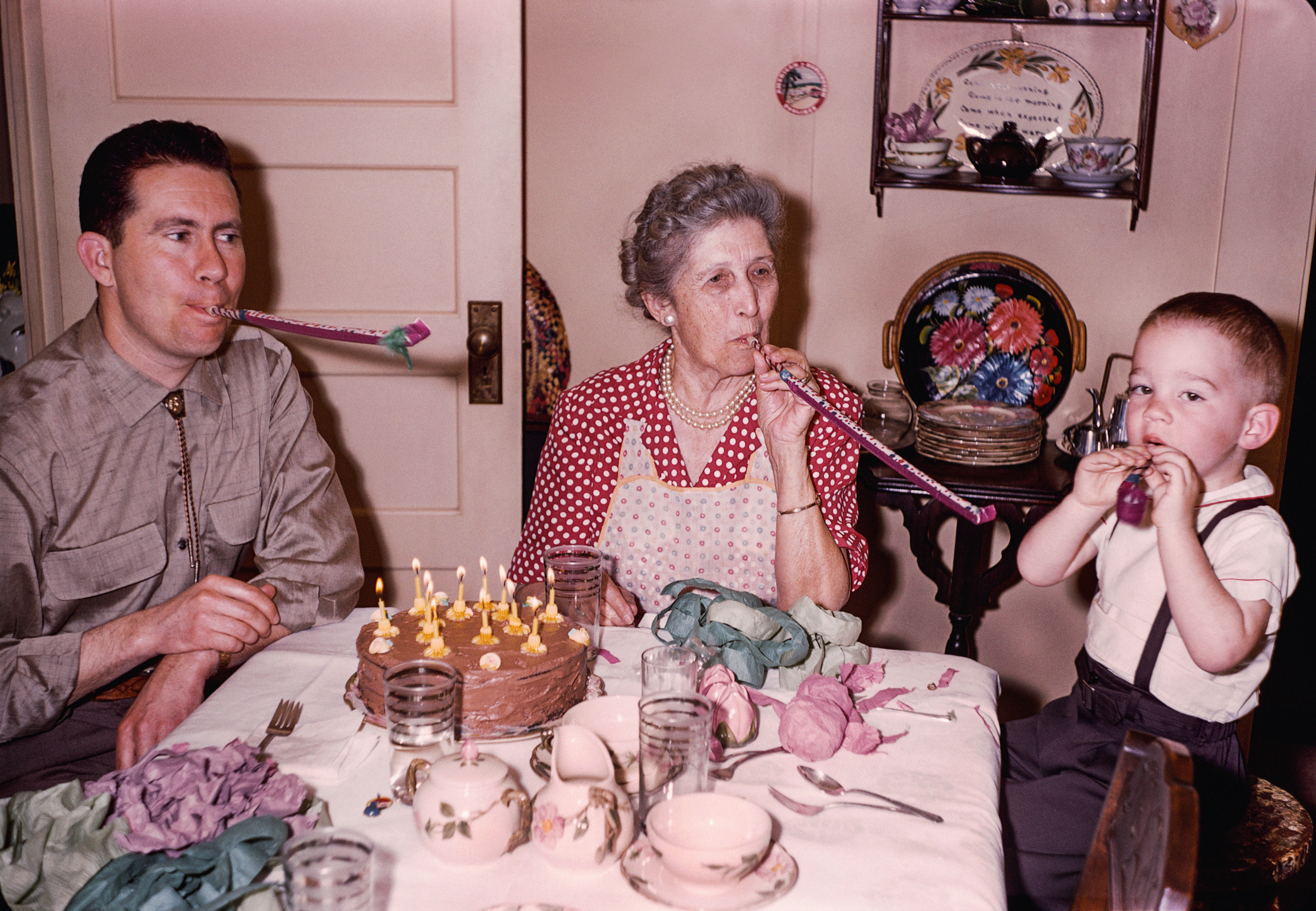 An older woman, a middle-aged man, and a young boy blow party horns at a table with a lit birthday cake. The room has vintage decor