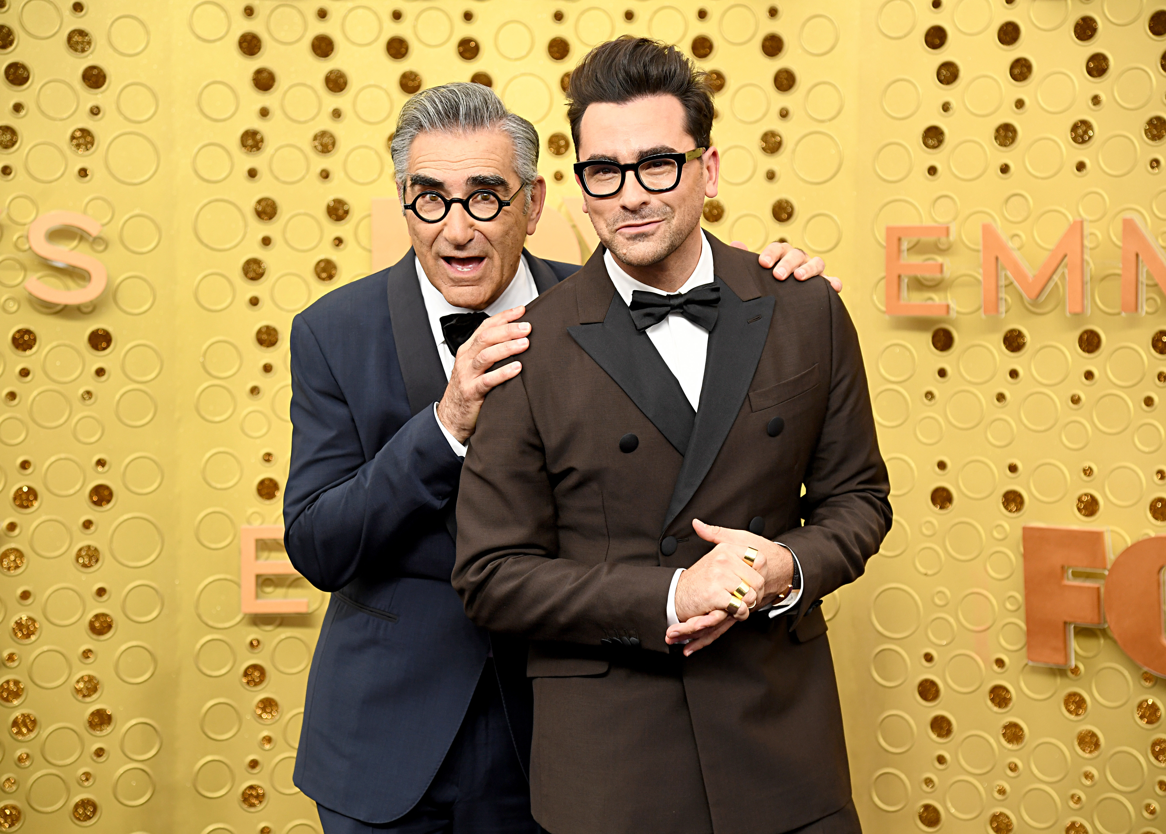 Eugene Levy and Dan Levy pose together on the red carpet. Eugene wears a navy suit and Dan wears a brown double-breasted suit with a bow tie