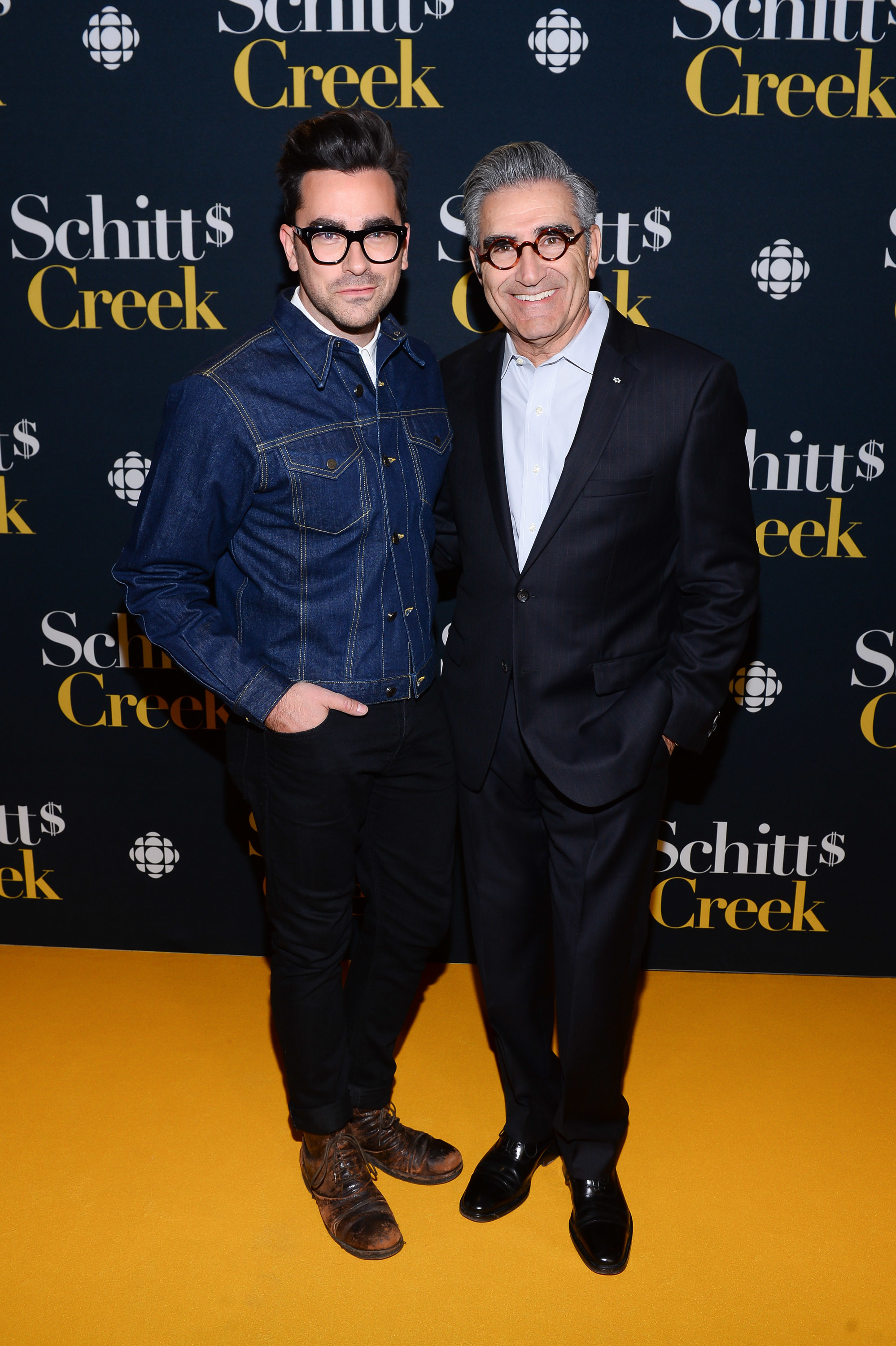 Dan Levy in a denim jacket and glasses, and Eugene Levy in a dark suit and glasses, pose together on the yellow carpet at the Schitt's Creek event