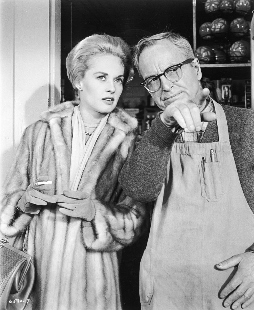 Tippi Hendren in a fur coat speaks with Alfred Hitchcock, who is wearing glasses and an apron, gesturing with his left hand
