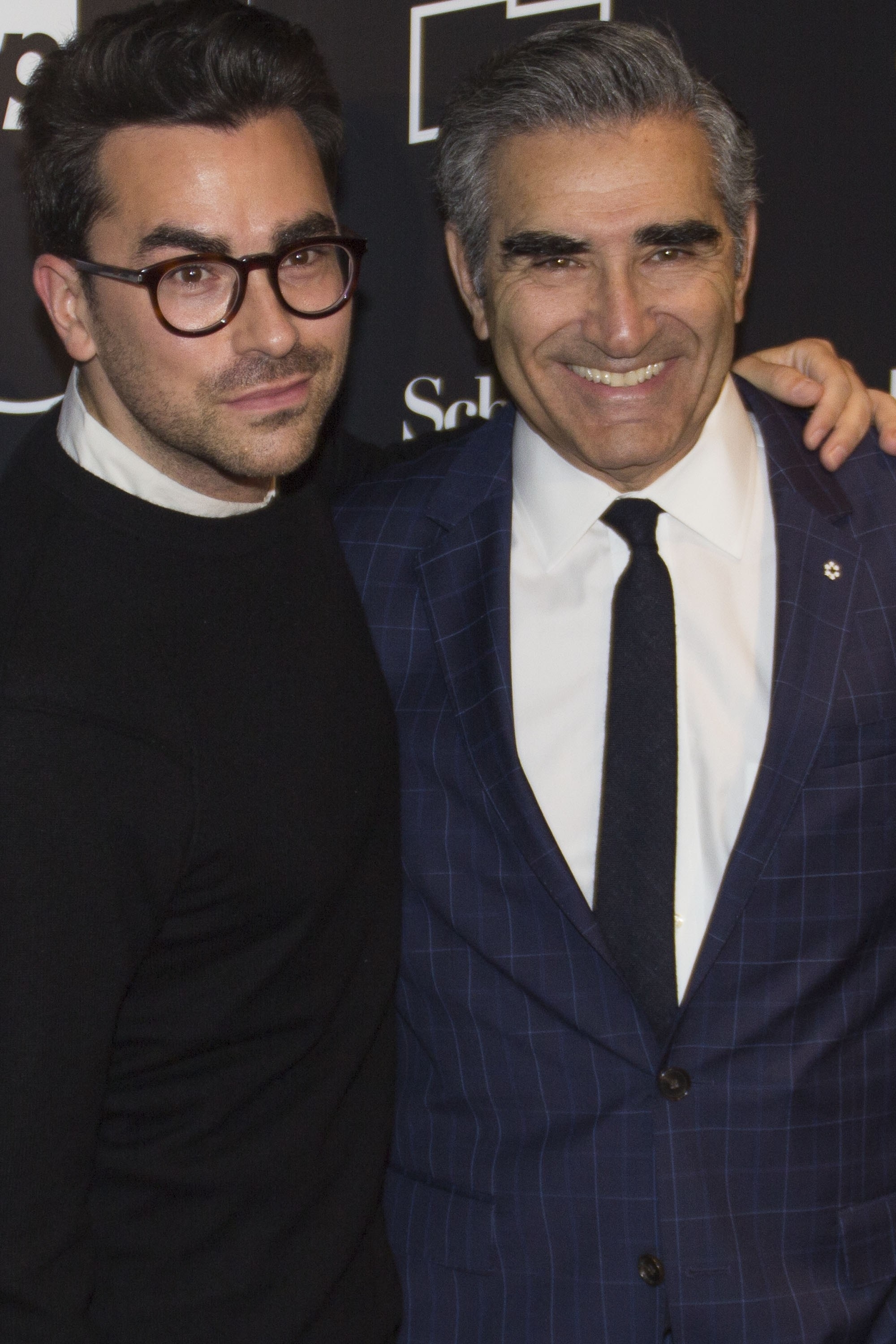 Dan Levy and Eugene Levy pose together at an event. Dan wears a black top and glasses, while Eugene wears a checkered suit with a white shirt and black tie