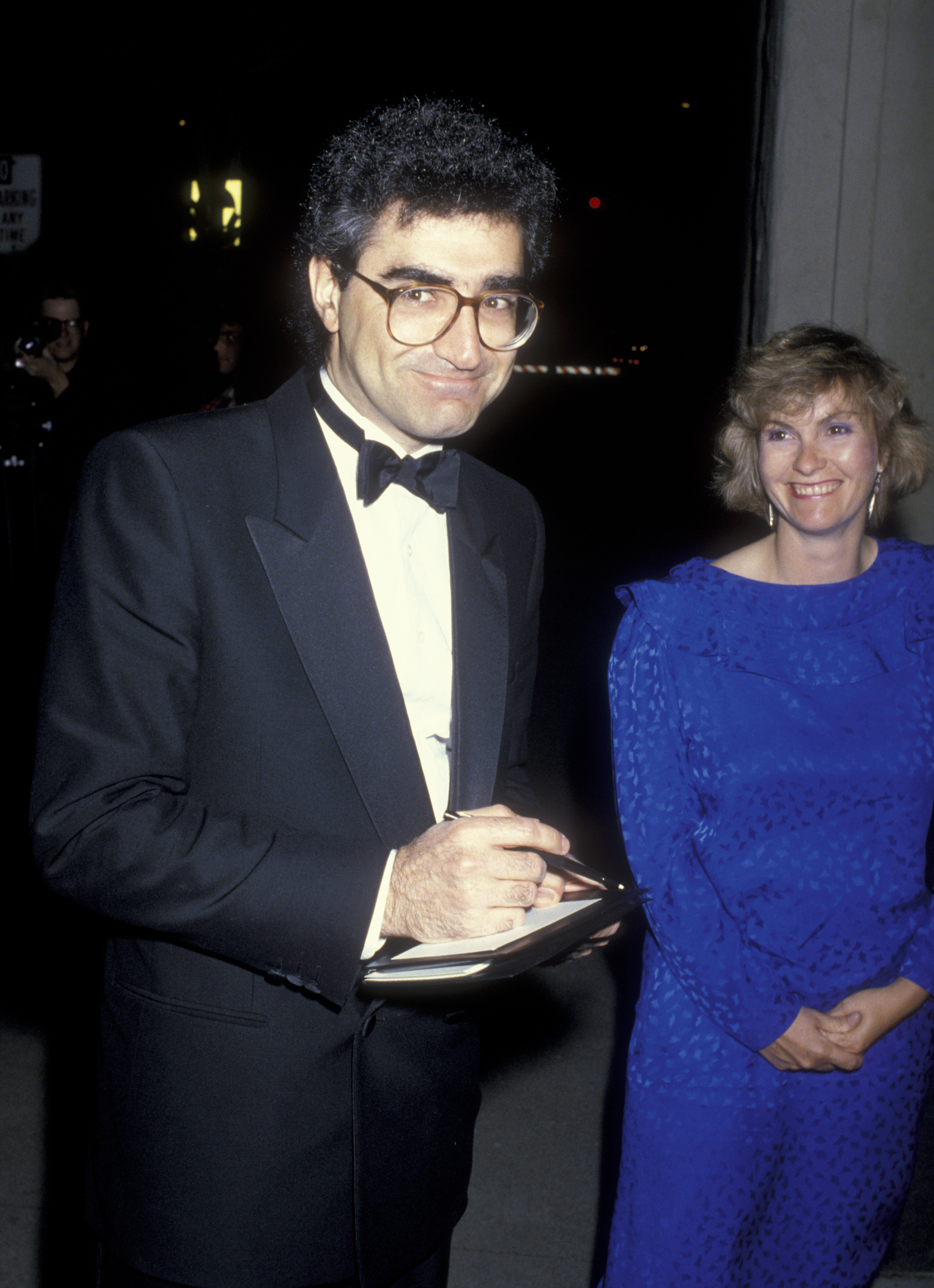 Eugene Levy in a black tuxedo stands next to Deborah Divine in a blue dress at a formal event. He is holding a pen and notepad