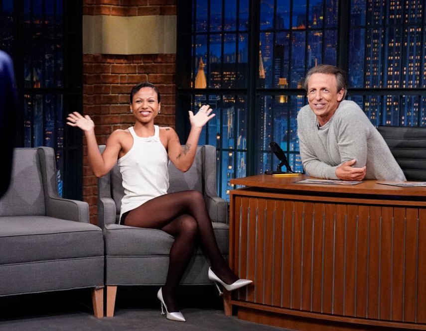 Myah'la sits on a late-night talk show set, smiling and waving her hands, while host Seth Meyers, dressed in a sweater, looks at her from behind his desk