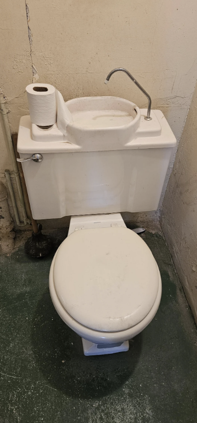 A small bathroom with a white toilet featuring a built-in sink on the tank, including a faucet, with a toilet paper roll on top and a plunger nearby
