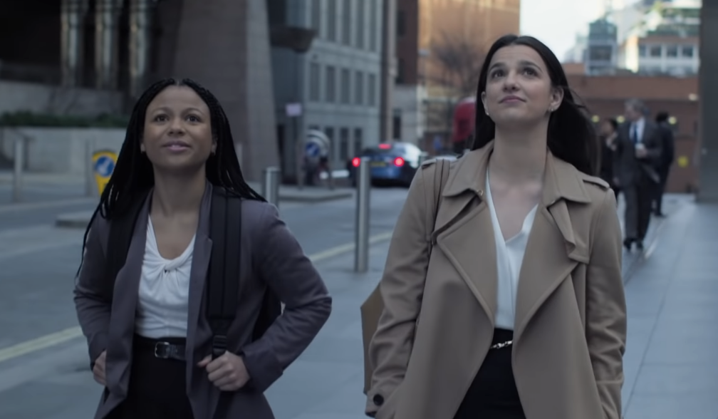 Two women, one with braids in a business suit, and one with straight hair in a beige coat over a white blouse, walking on a city street