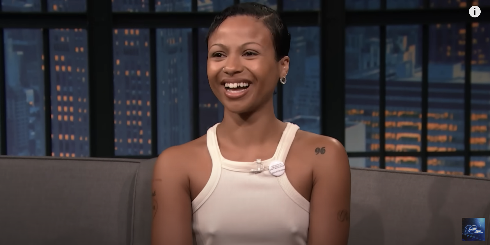 A person with short hair smiles while sitting on a talk show set. In the background, city buildings are visible at night. The person wears a sleeveless top