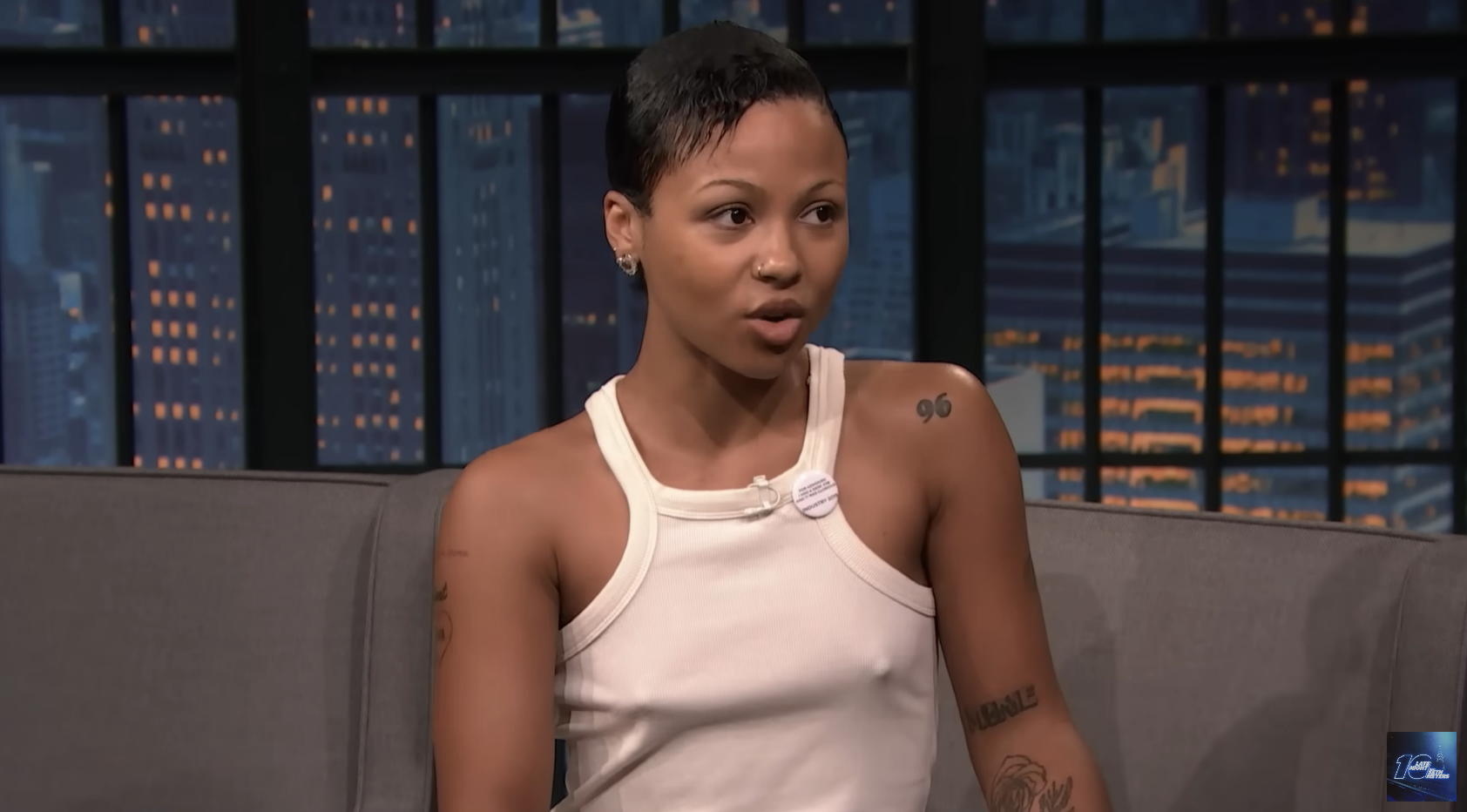 A woman with short hair and tattoos, wearing a sleeveless top, speaks during a late-night talk show interview