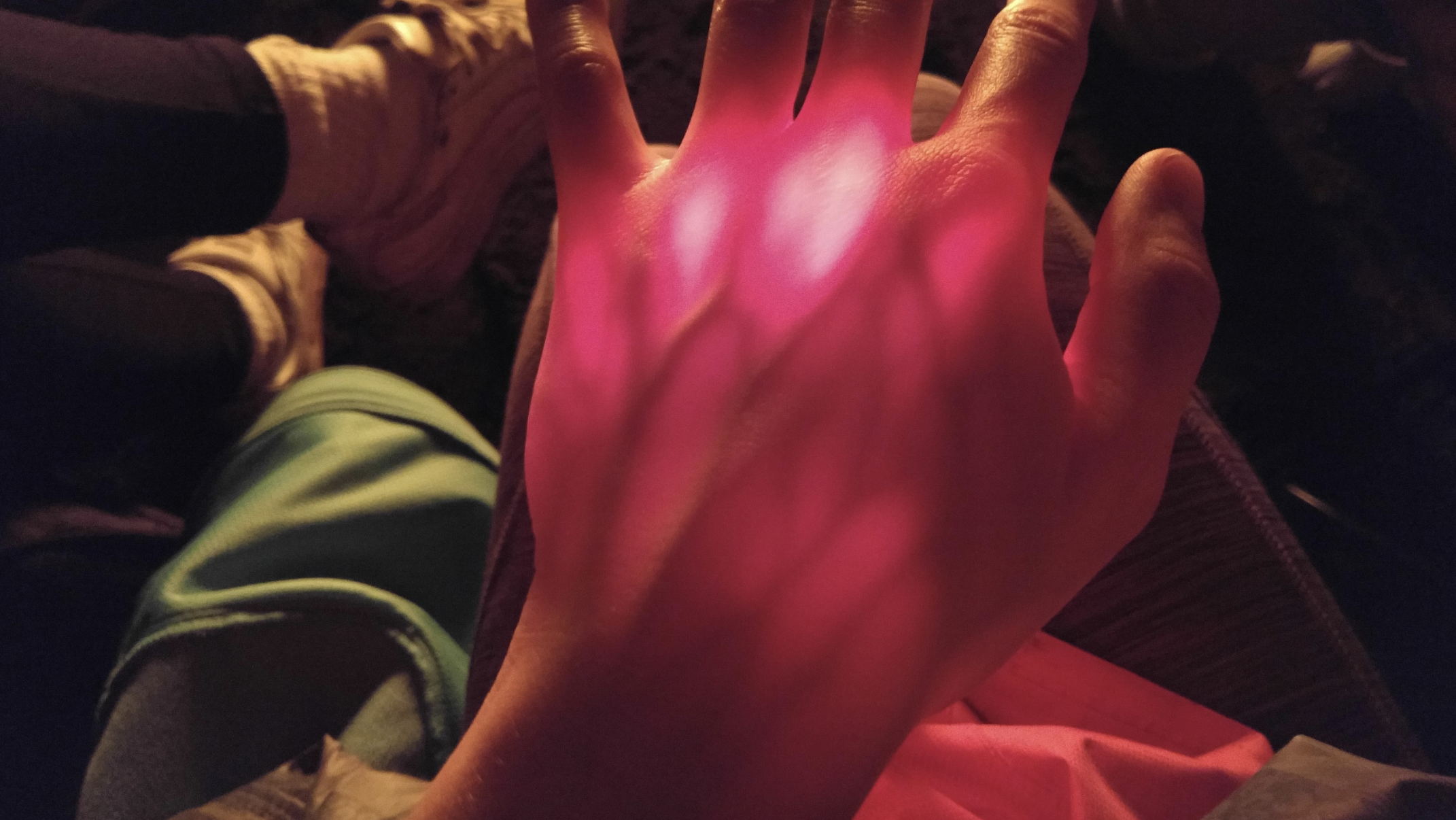 A hand illuminated with pink light is shown close up, with people in the background