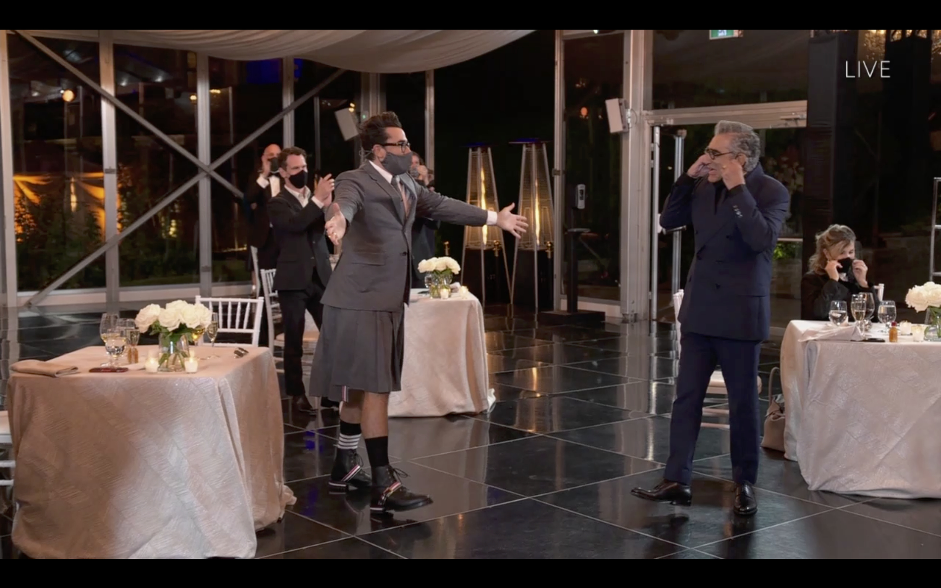 Dan Levy, in a kilt with suit jacket, embraces Eugene Levy, in a suit, during an awards ceremony in a stylish, well-lit venue. Other attendees are seated at tables