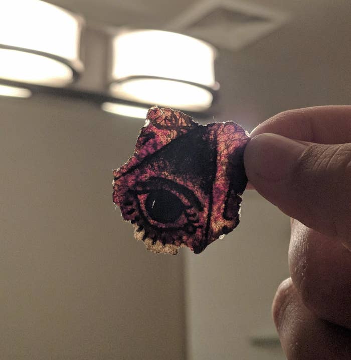 A hand holds a piece of artwork resembling a detailed eye on a small, irregularly shaped material. The background is out of focus, showing ceiling lights