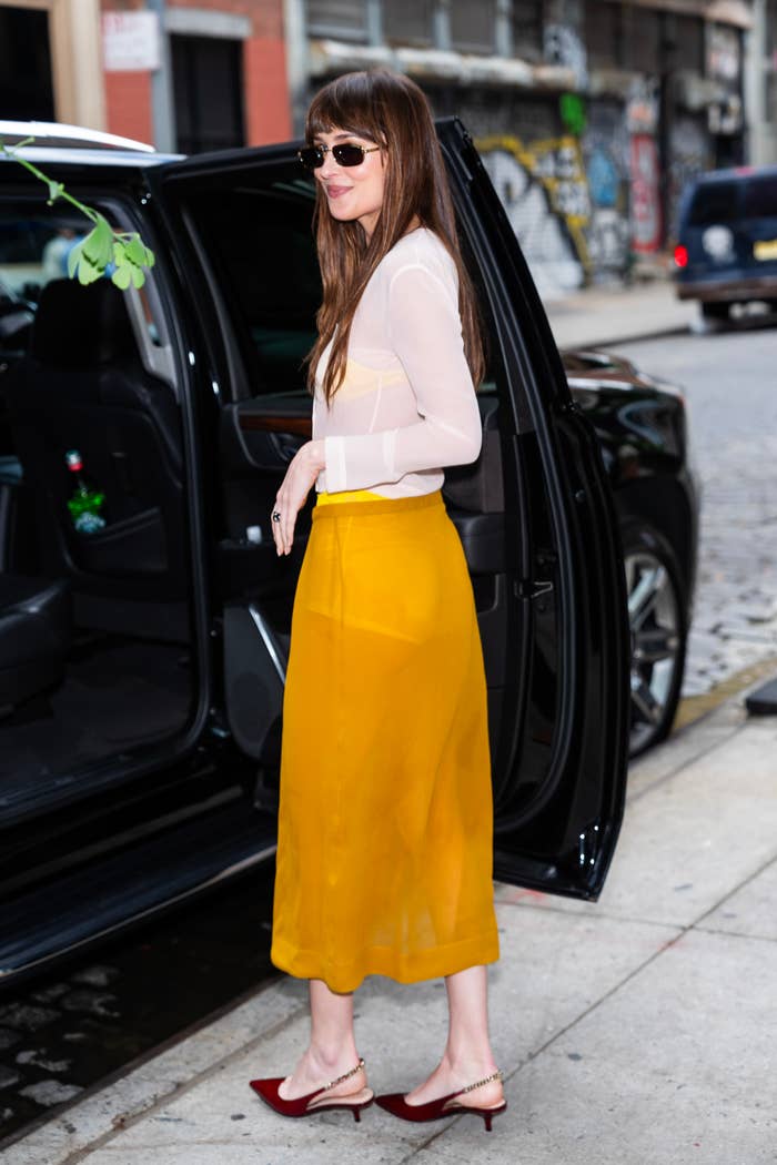 Dakota Johnson steps out of a car wearing a sheer blouse, bright yellow skirt, and red heels, with her hair down and sunglasses on, in a city street environment