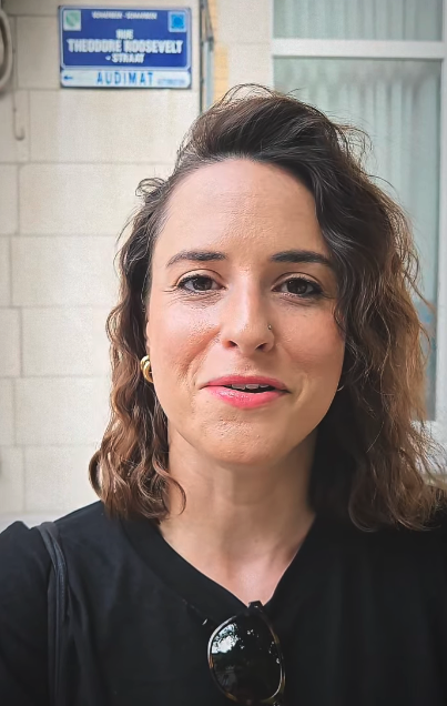 Rachel with shoulder-length wavy hair, wearing a black shirt, standing in front of a sign reading "Rue Theodore Roosevelt, Audimat."