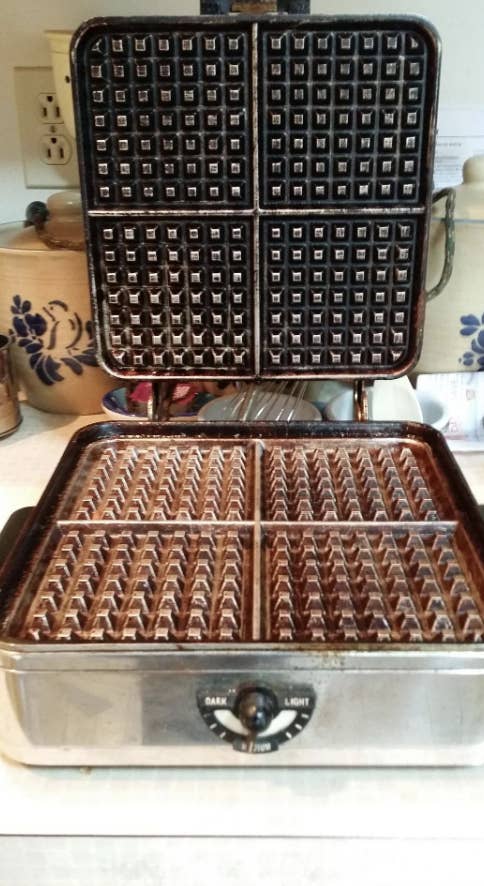 Open waffle iron on a kitchen counter with visible control knob labeled "dark" and "light." Various kitchen items are in the background
