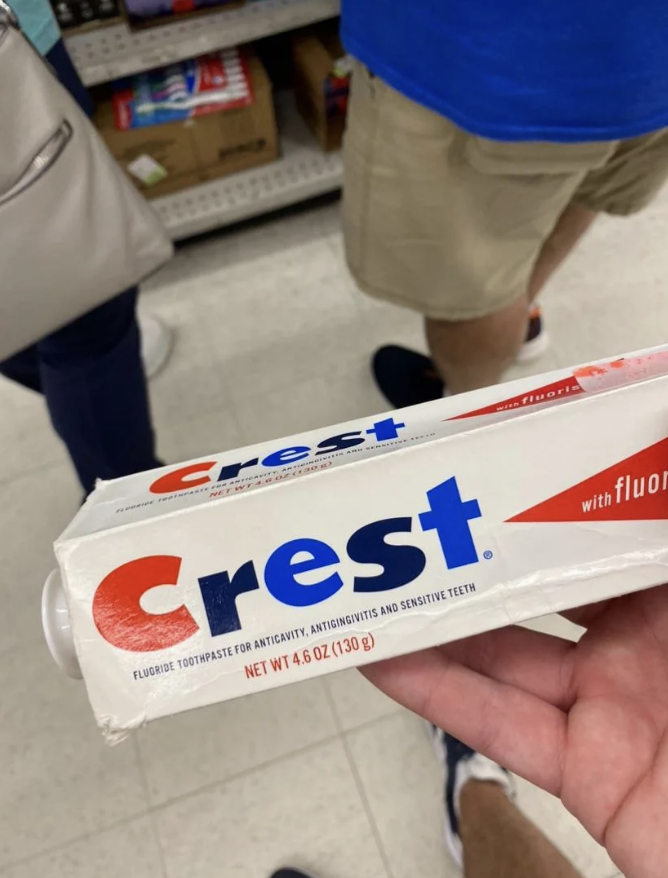 A person holds a 4.6 oz tube of Crest toothpaste in a store. Text on the toothpaste reads "Fluoride toothpaste for anticavity, antigingivitis, and sensitive teeth."