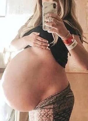 A person with long hair takes a mirror selfie, showing a large pregnant belly and wearing a black top and patterned pants